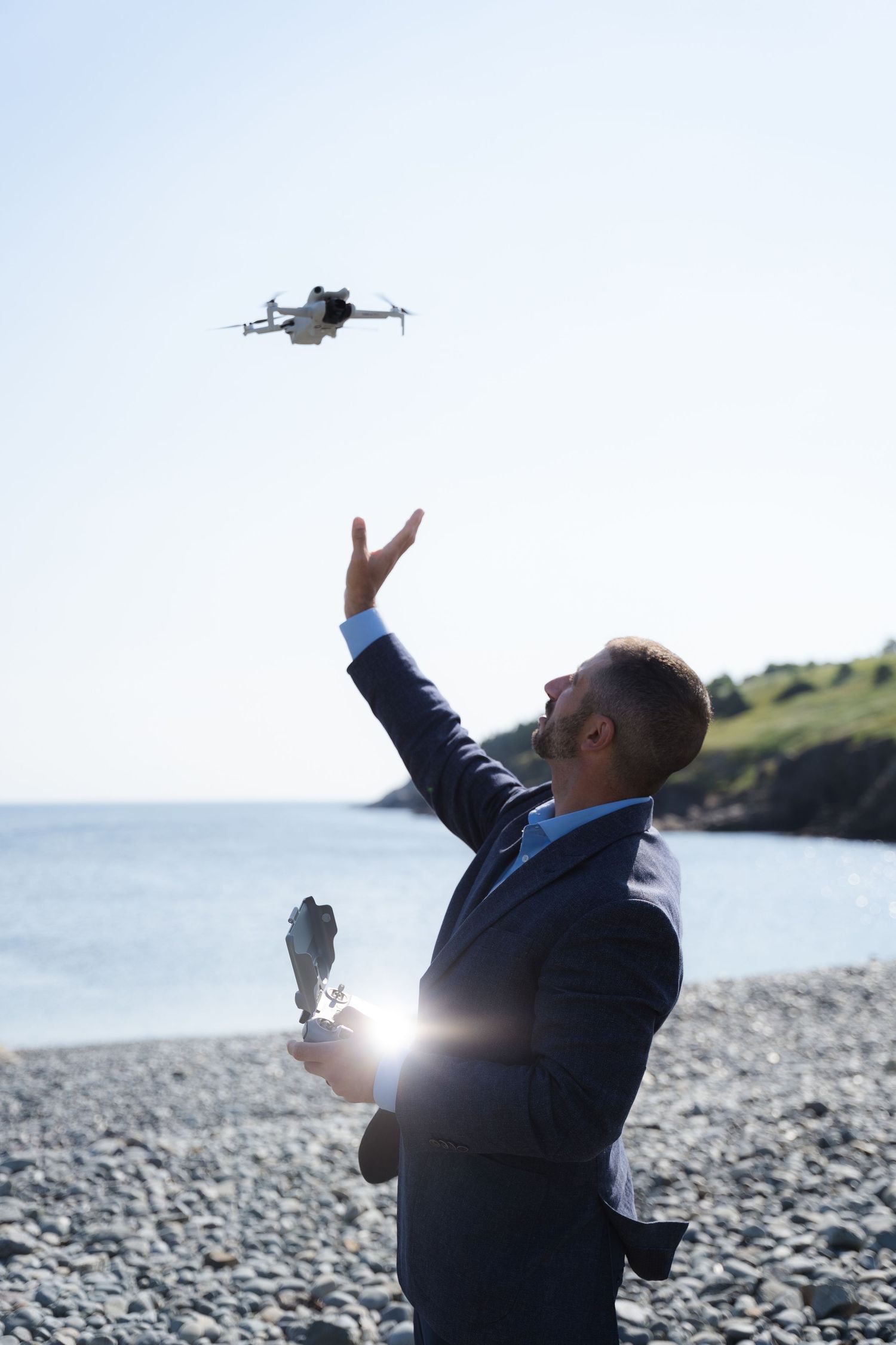 Businessman in suit launching a drone on a rocky beach with ocean and green hills in background.