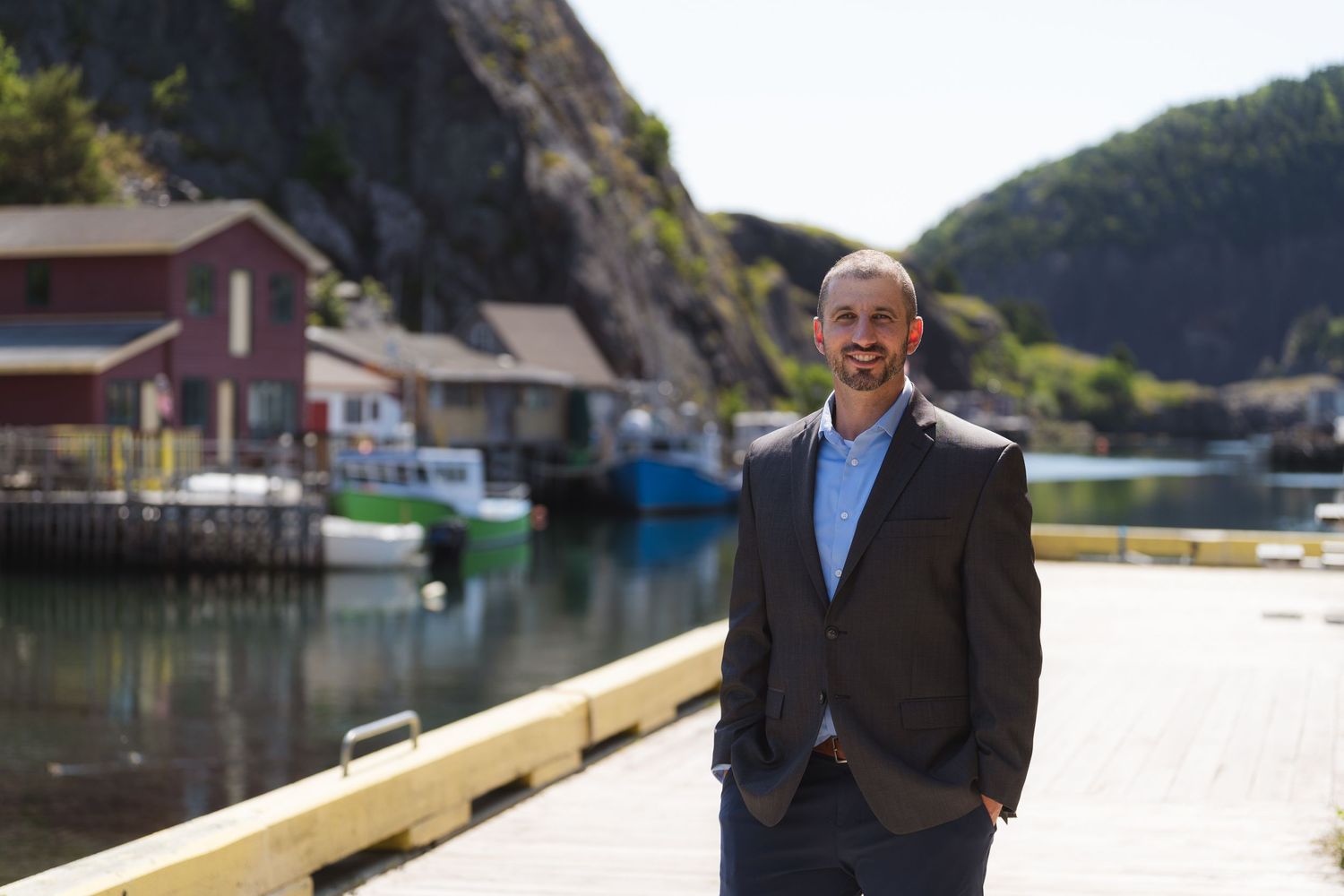 Man in suit smiling on a waterfront dock with colorful boats and rocky hillside village in background.