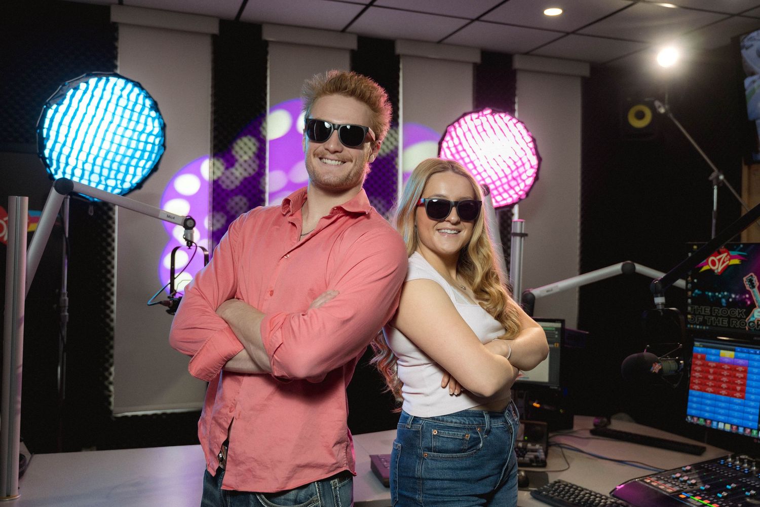 Two smiling DJs wearing sunglasses pose back-to-back in a colorful radio studio with stage lights.