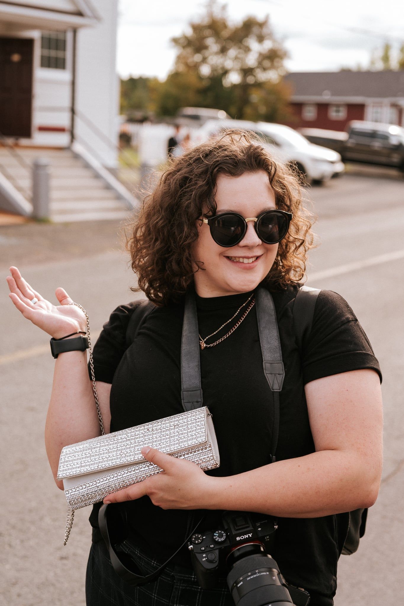 A photographer with curly hair and sunglasses smiling while holding camera gear.