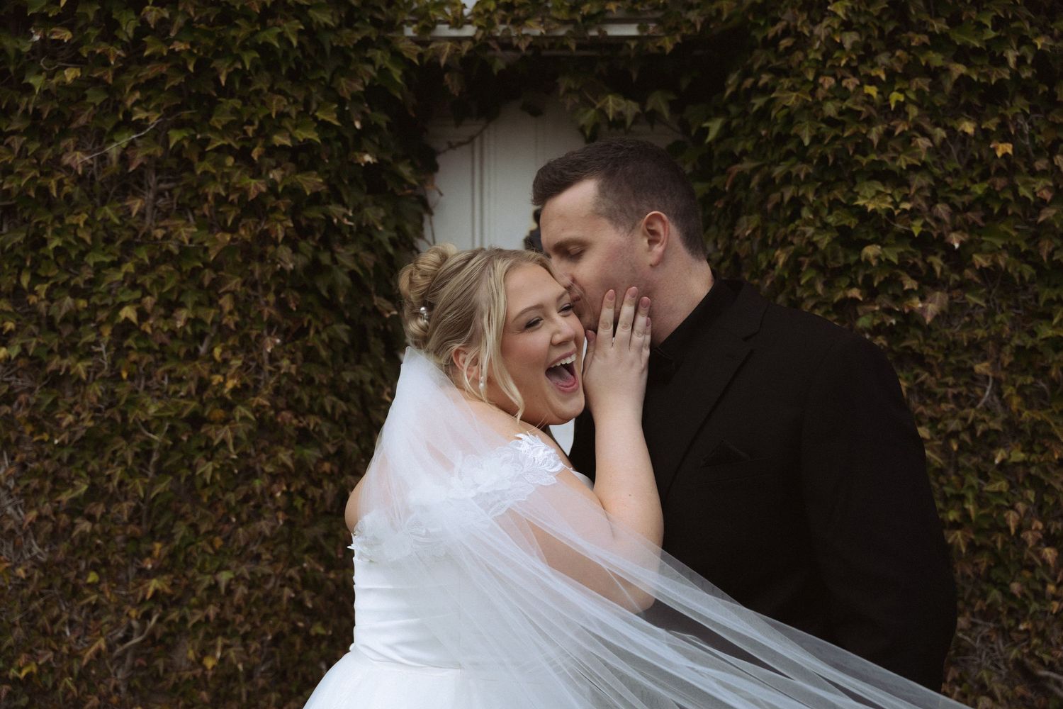 A couple in wedding attire shares an intimate moment against a lush green hedge background.