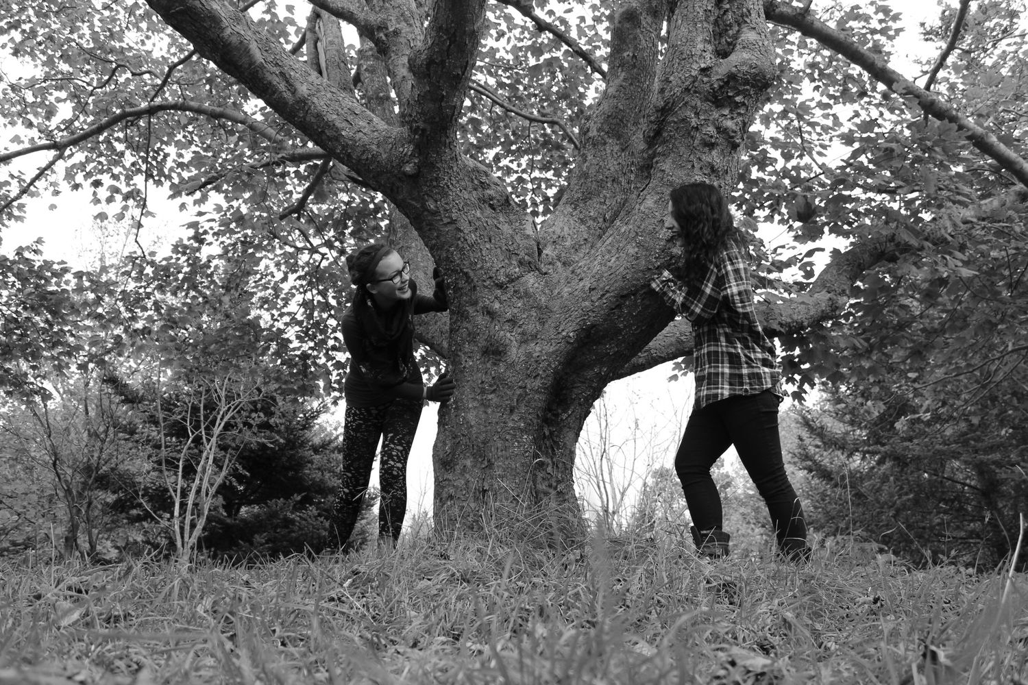 Two people playfully pose by a large tree trunk in a black and white photograph.