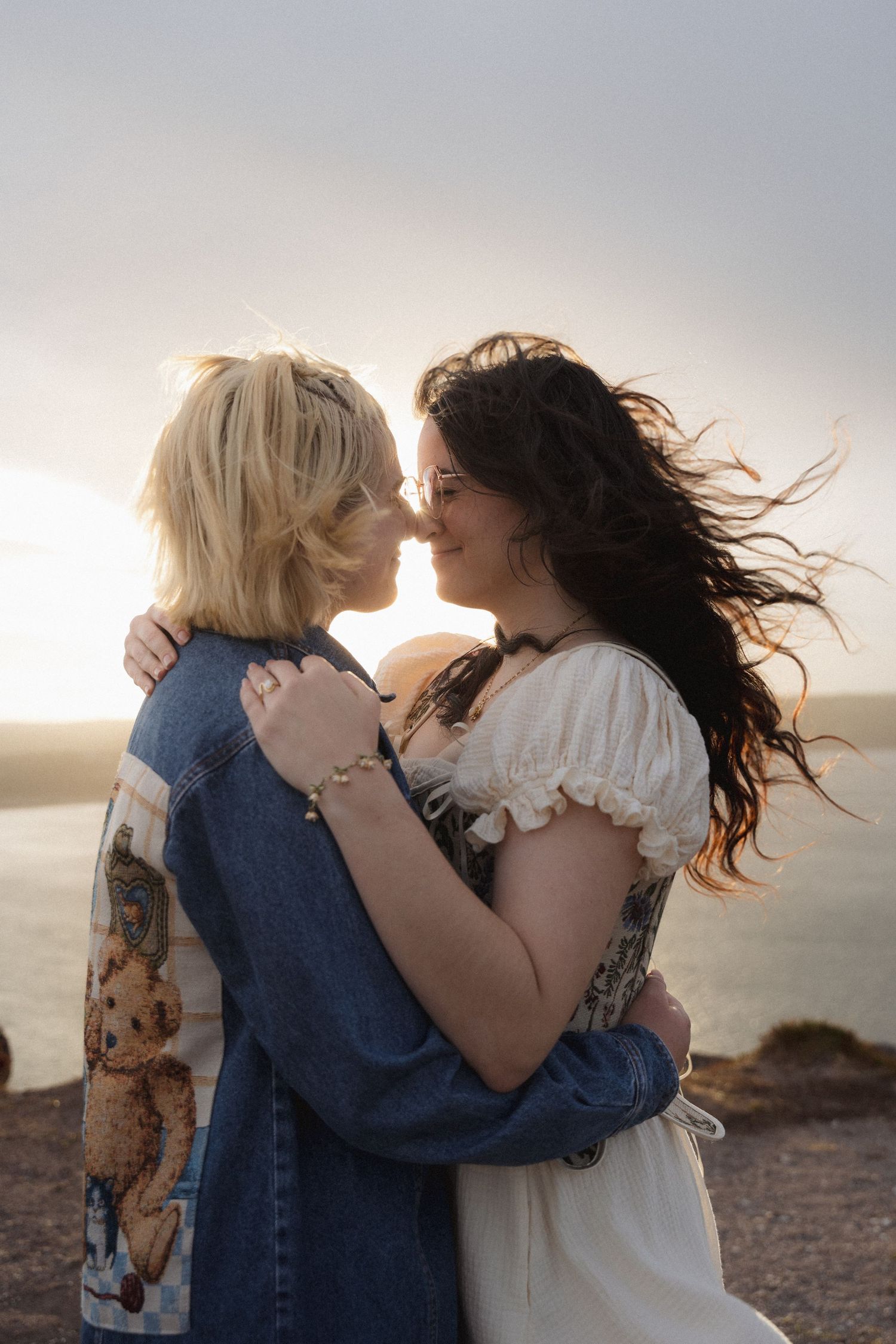 Two people embrace and dance together at sunset on a desert landscape while their hair blows in the wind.