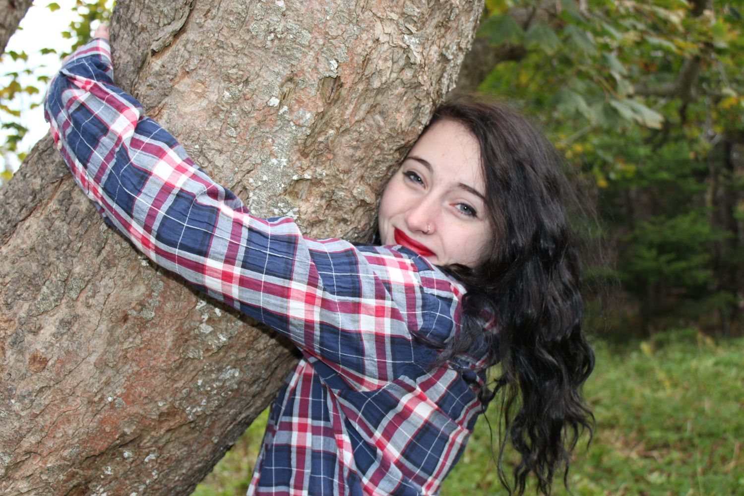 Person in plaid shirt poses playfully against a large tree trunk in a forest setting.