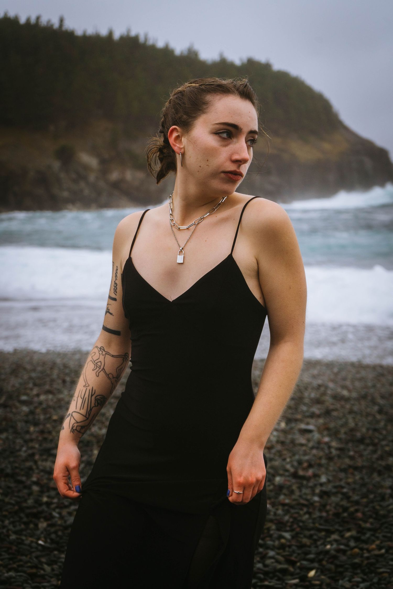 Person in black dress walks along rocky beach with waves and cliffs in background.