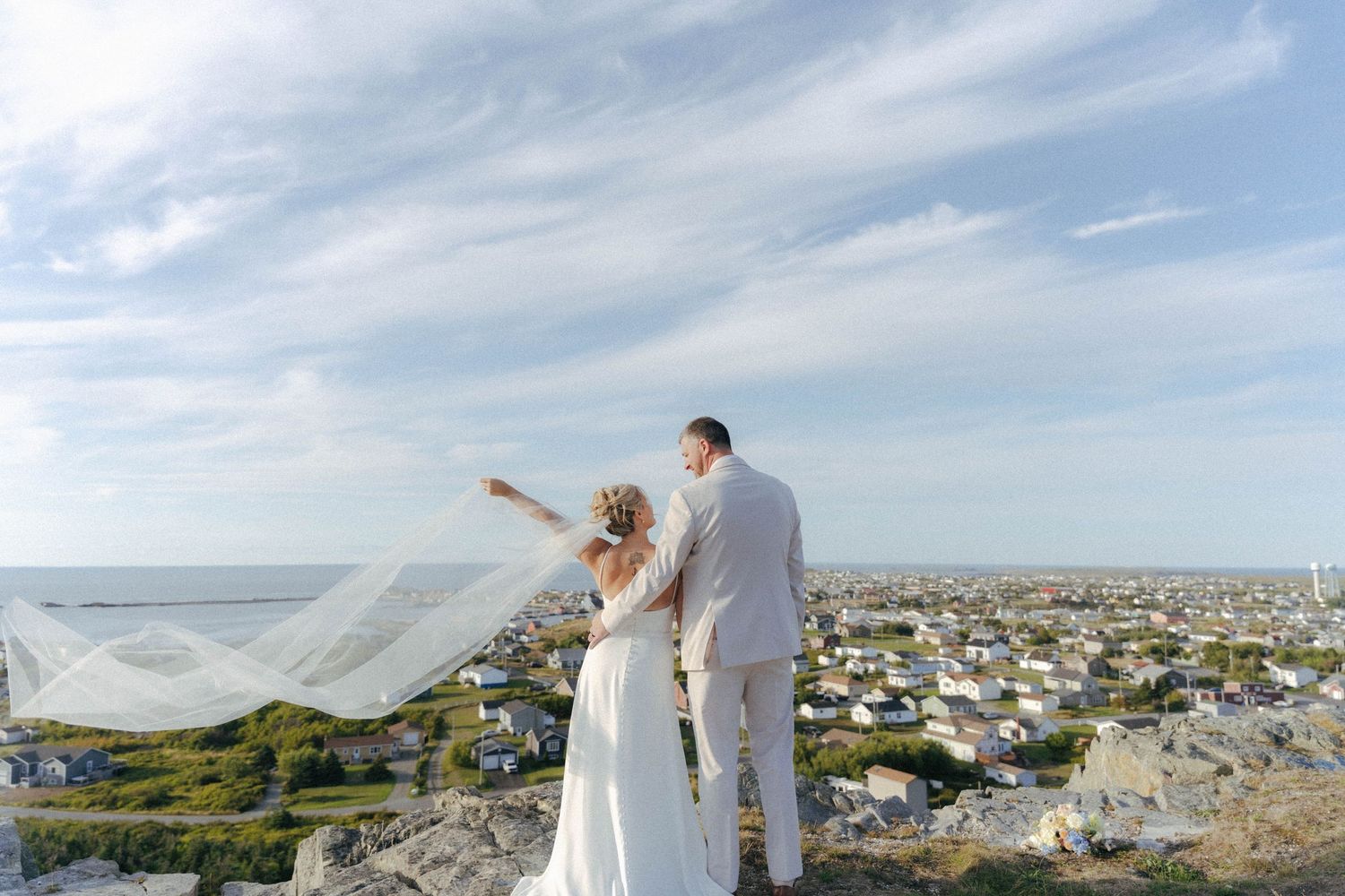 A bride and groom stand on a hilltop overlooking a coastal town as her veil flows dramatically in the wind.