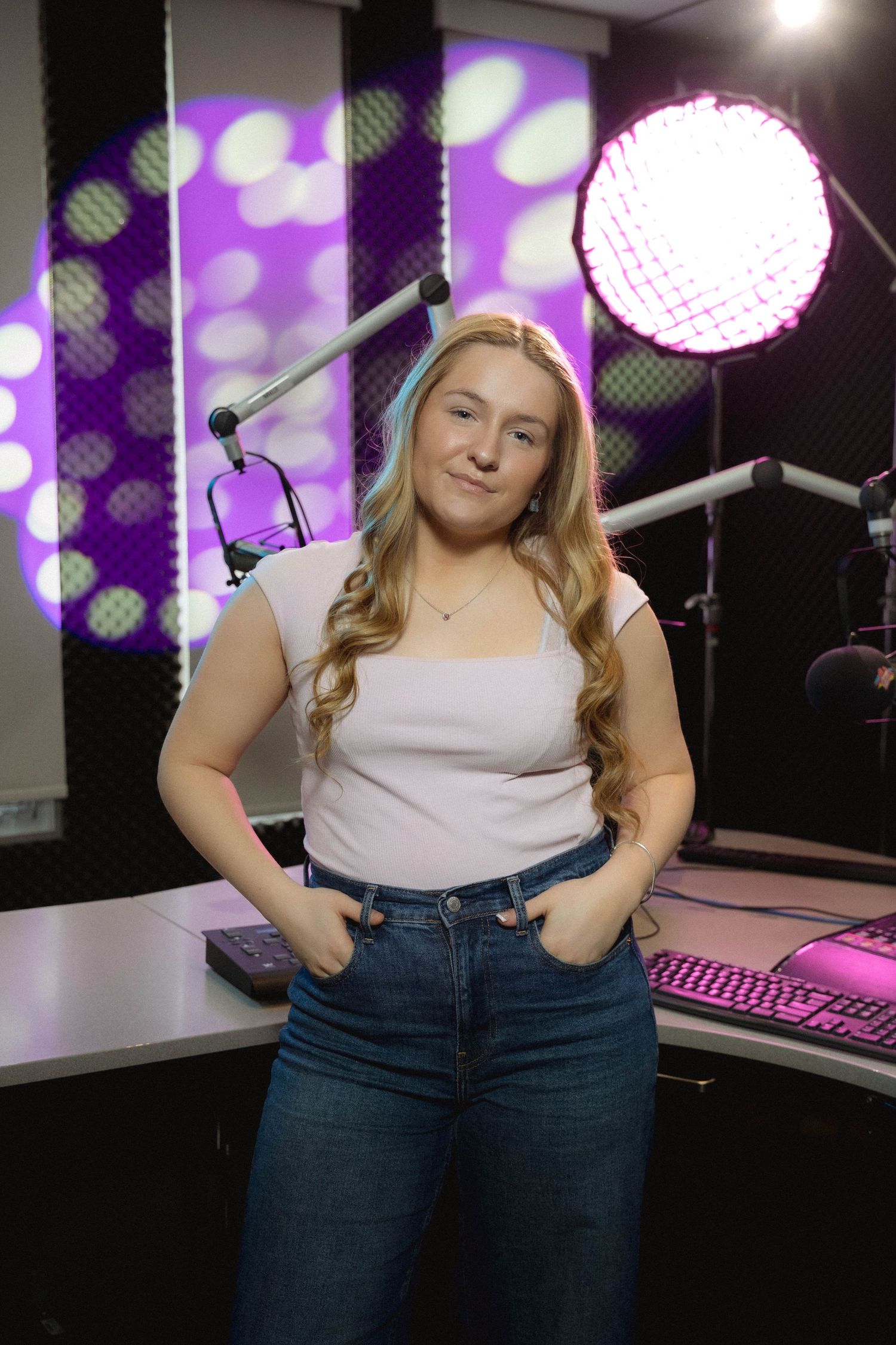 Young blonde woman poses confidently in a radio studio with purple lighting and microphones in the background.