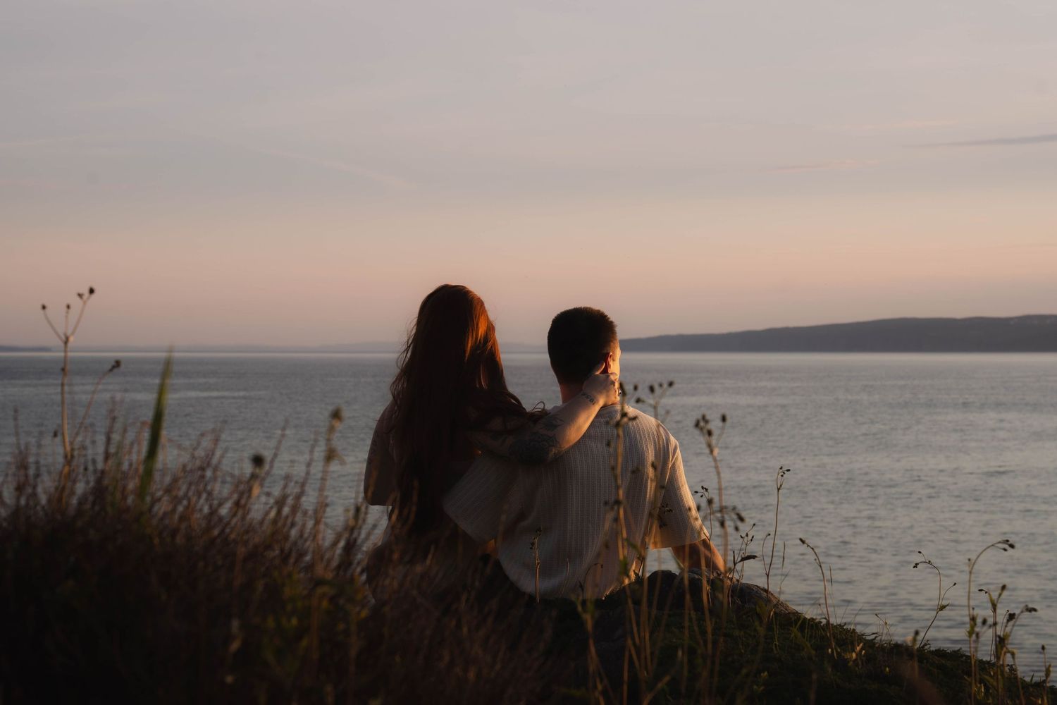 Silhouettes of couple by the ocean during sunset with coastline in background.