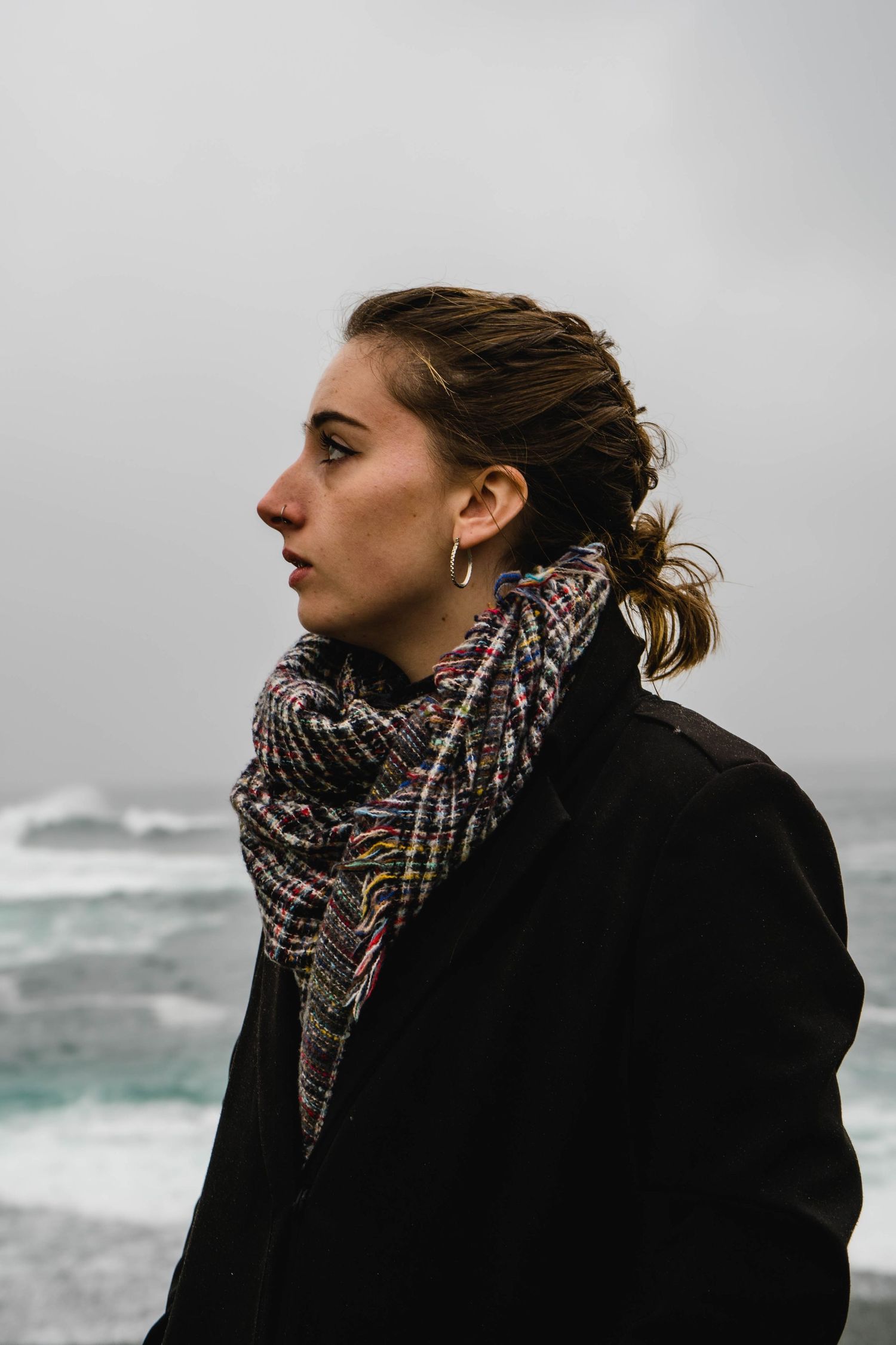 Person in black coat and patterned scarf gazes at stormy ocean waves.