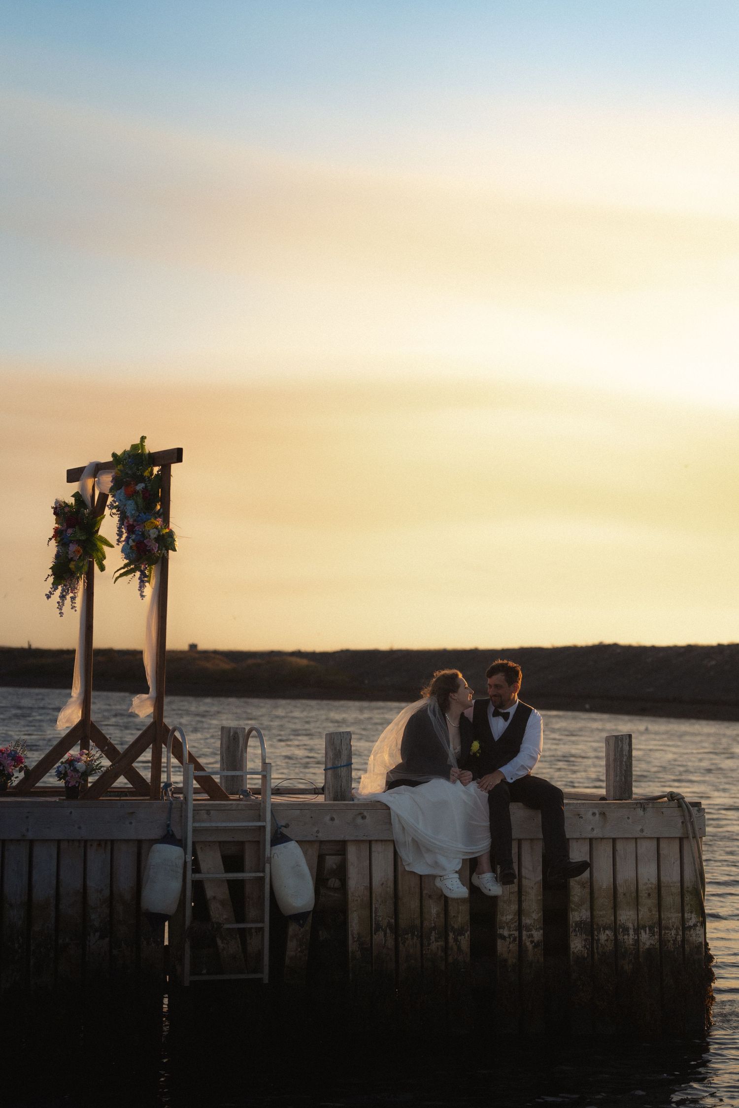 A romantic sunset silhouette of a couple at a waterfront dock decorated with floral arrangements.