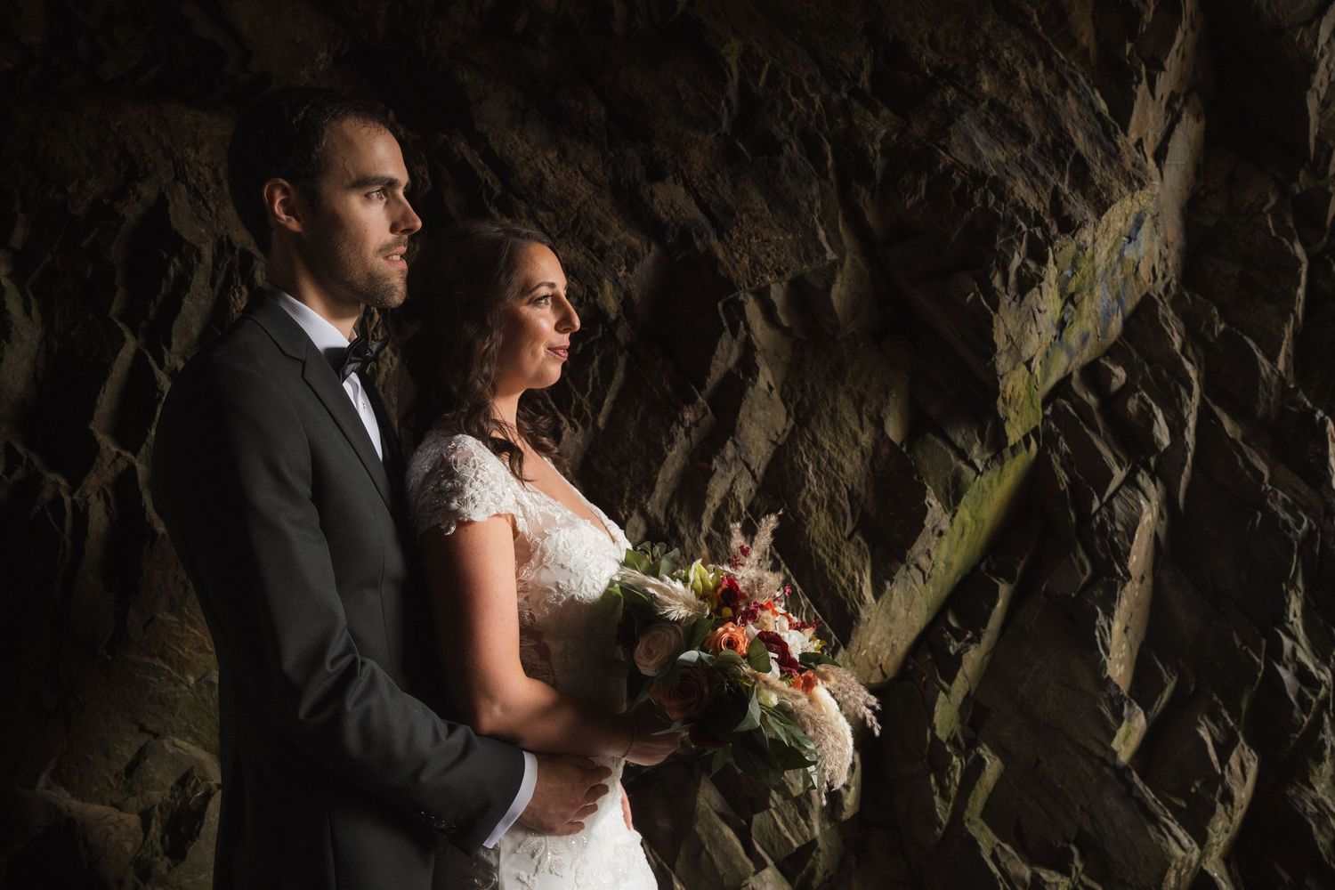 Wedding couple standing in dramatic cave setting with bouquet.
