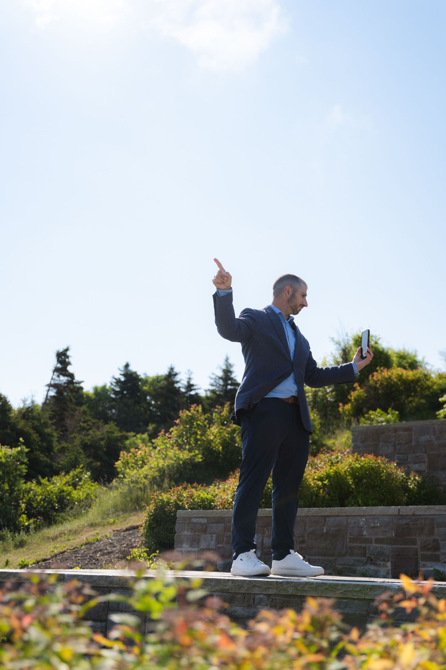 Businessman in suit pointing upward while holding tablet outdoors in sunny garden setting.