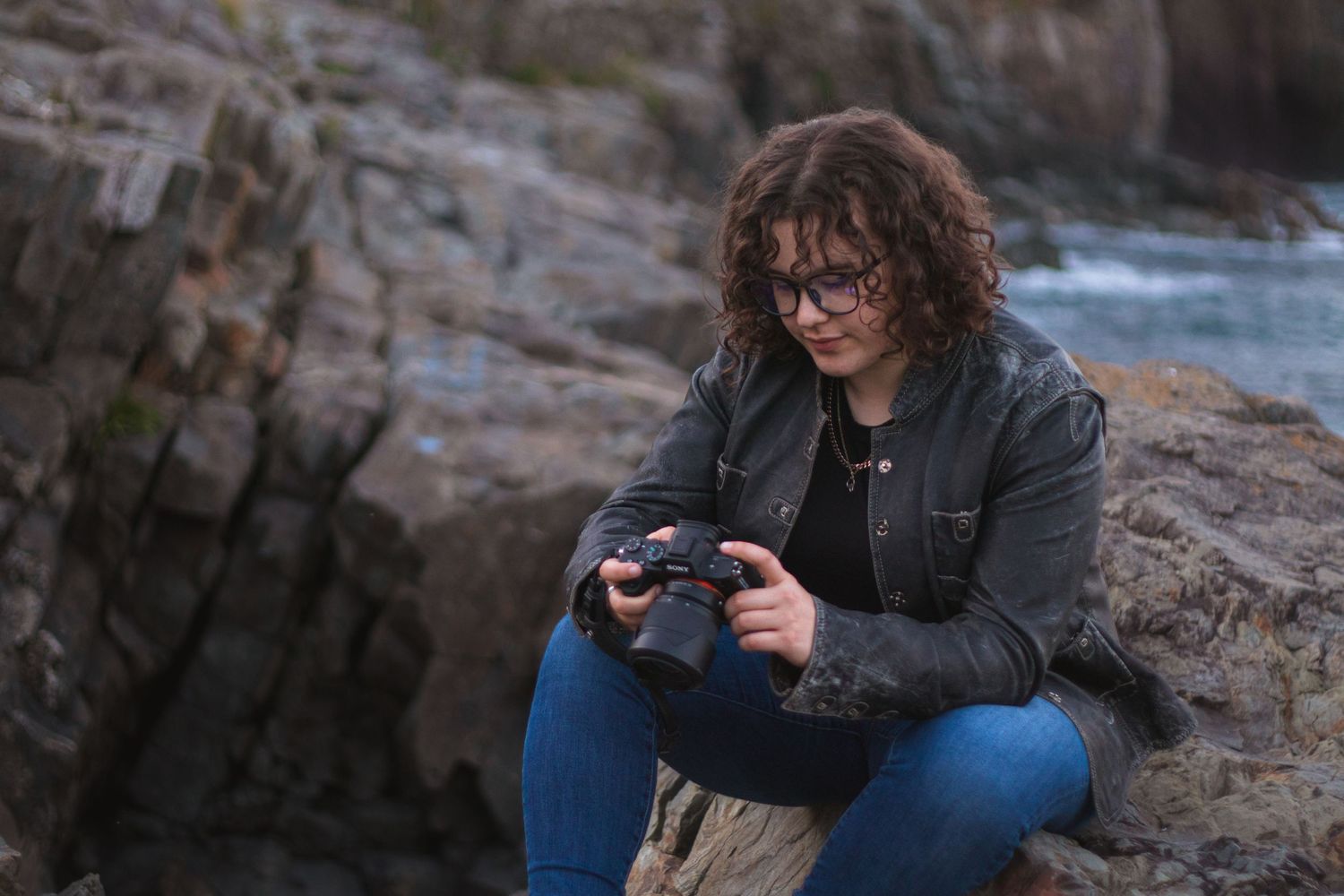 A photographer reviews images on their camera while sitting on coastal rocks.