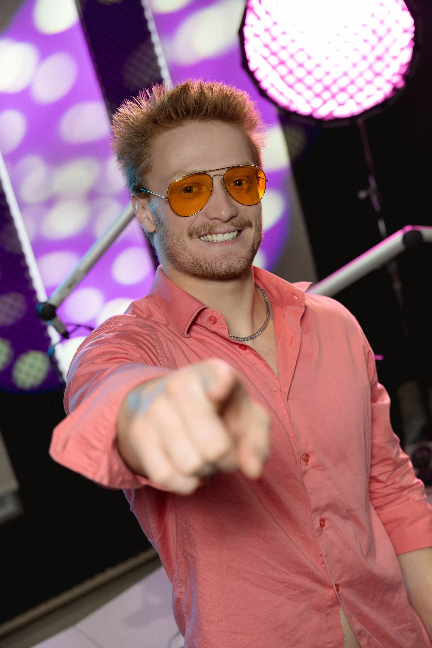 Smiling young man with orange aviator sunglasses and pink shirt pointing at camera in front of purple disco lights.