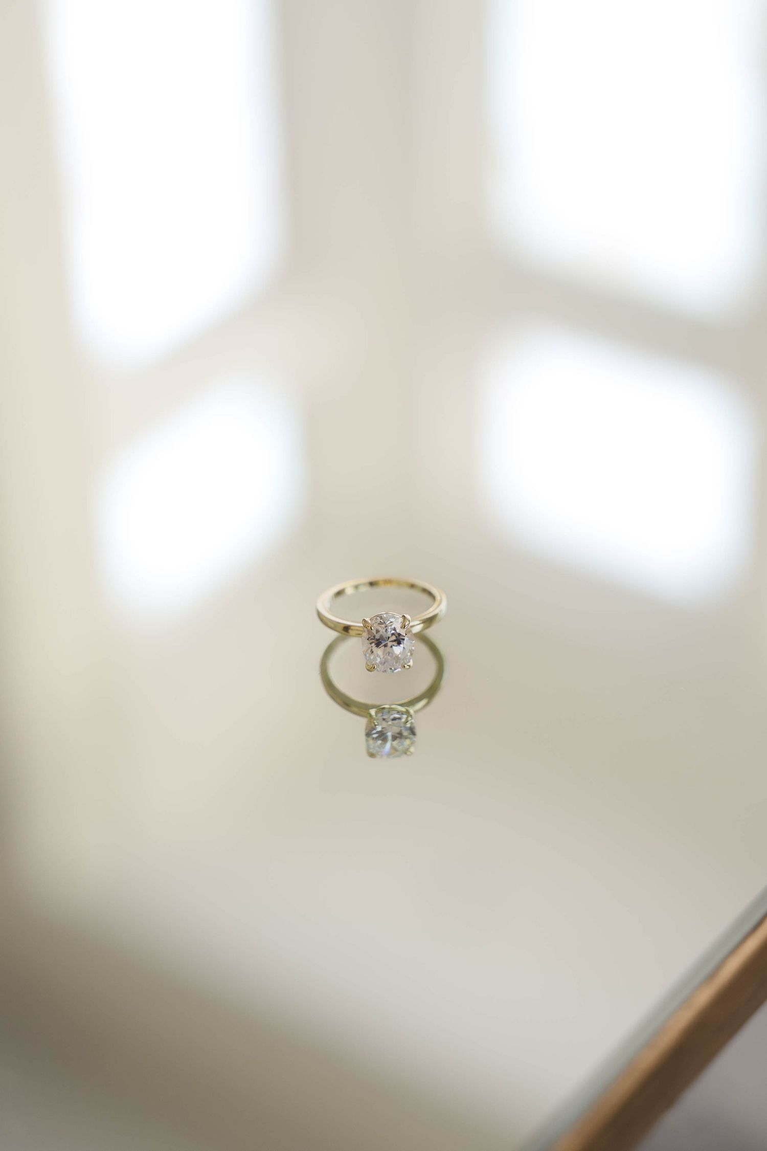 Gold solitaire diamond engagement ring with reflection on white marble surface in soft natural light.