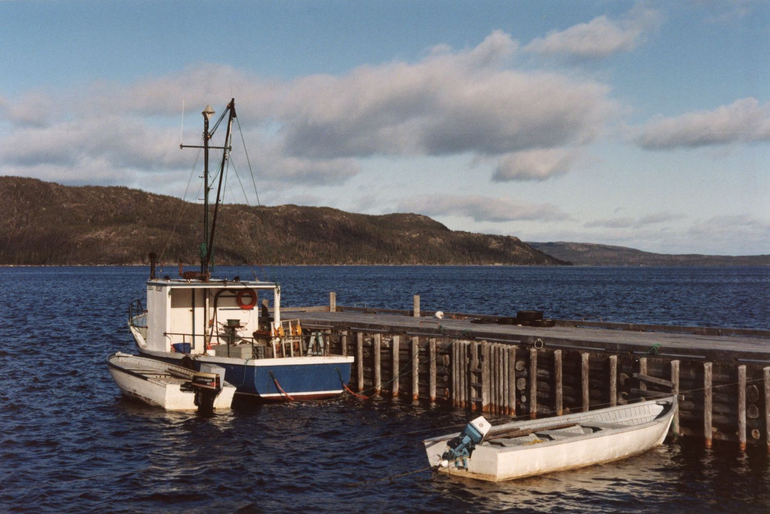 Panoramic view of a wooden dock extending into a serene lake with boats and mountains in the background.