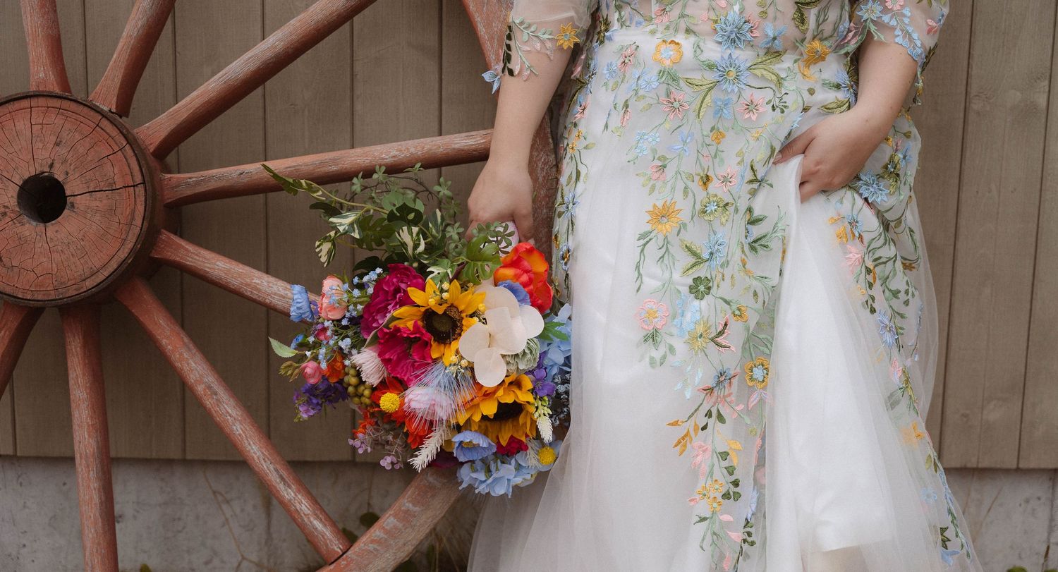 A floral wedding dress is showcased in a rustic barn setting with wooden wagon wheels and colorful flower bouquets.