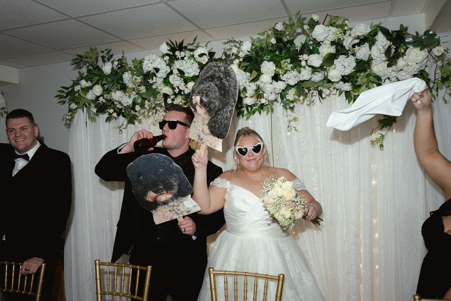 Wedding party members wearing sunglasses pose playfully during reception under white floral arch decor.
