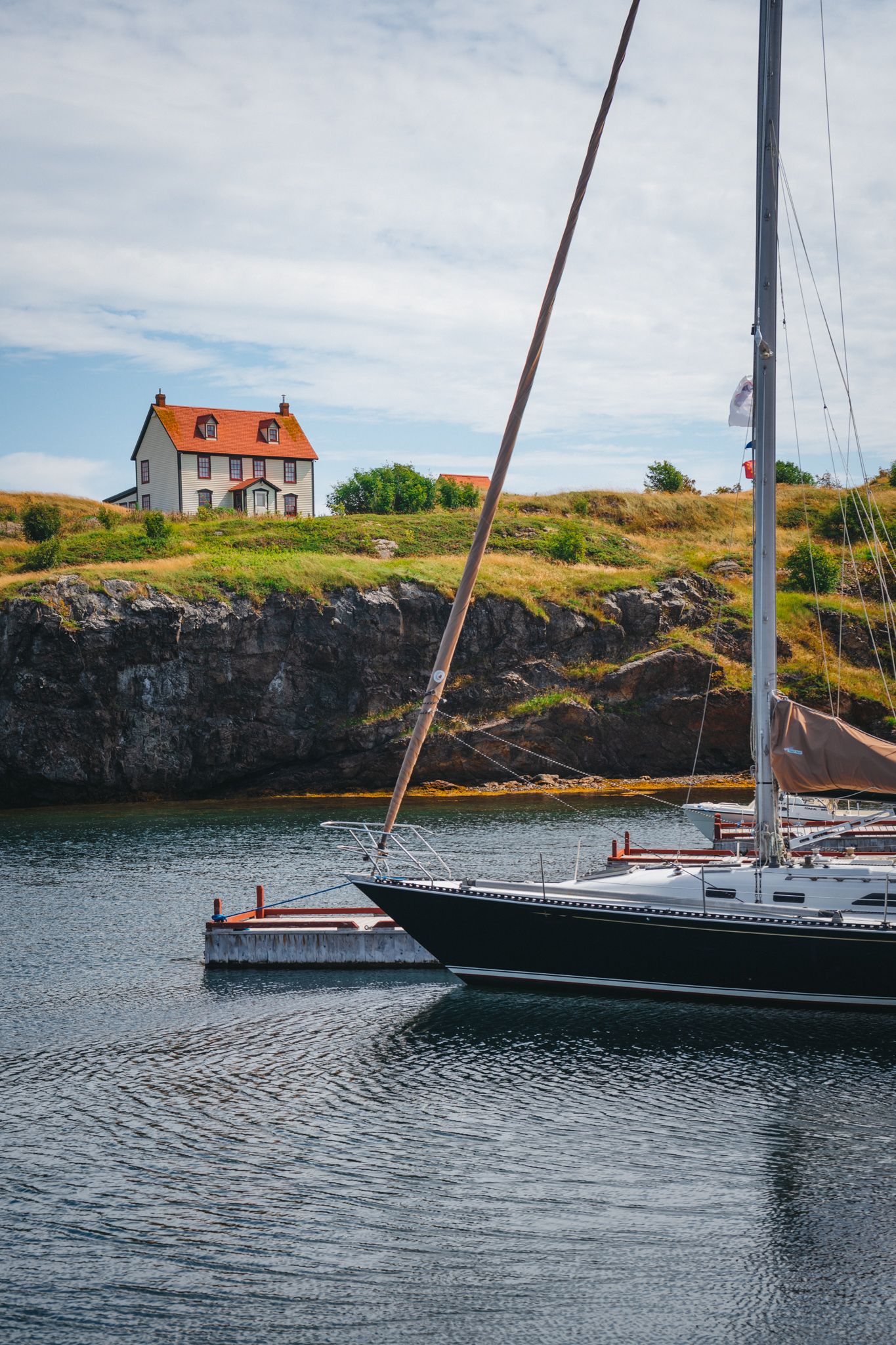 A sailboat moored near a rustic orange house perched on rocky coastal cliffs.