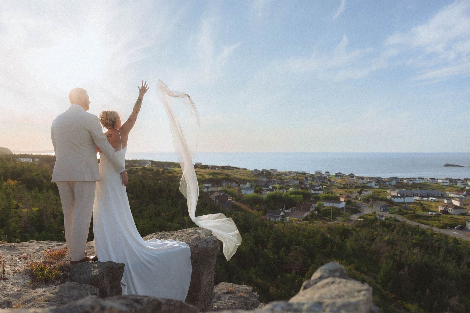 Couple in wedding attire stands on coastal cliffs overlooking the ocean during golden hour.