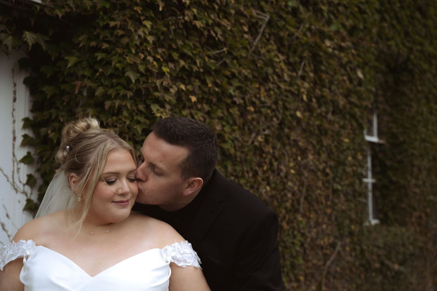 Romantic outdoor wedding scene with bride and groom embracing against an ivy-covered wall.