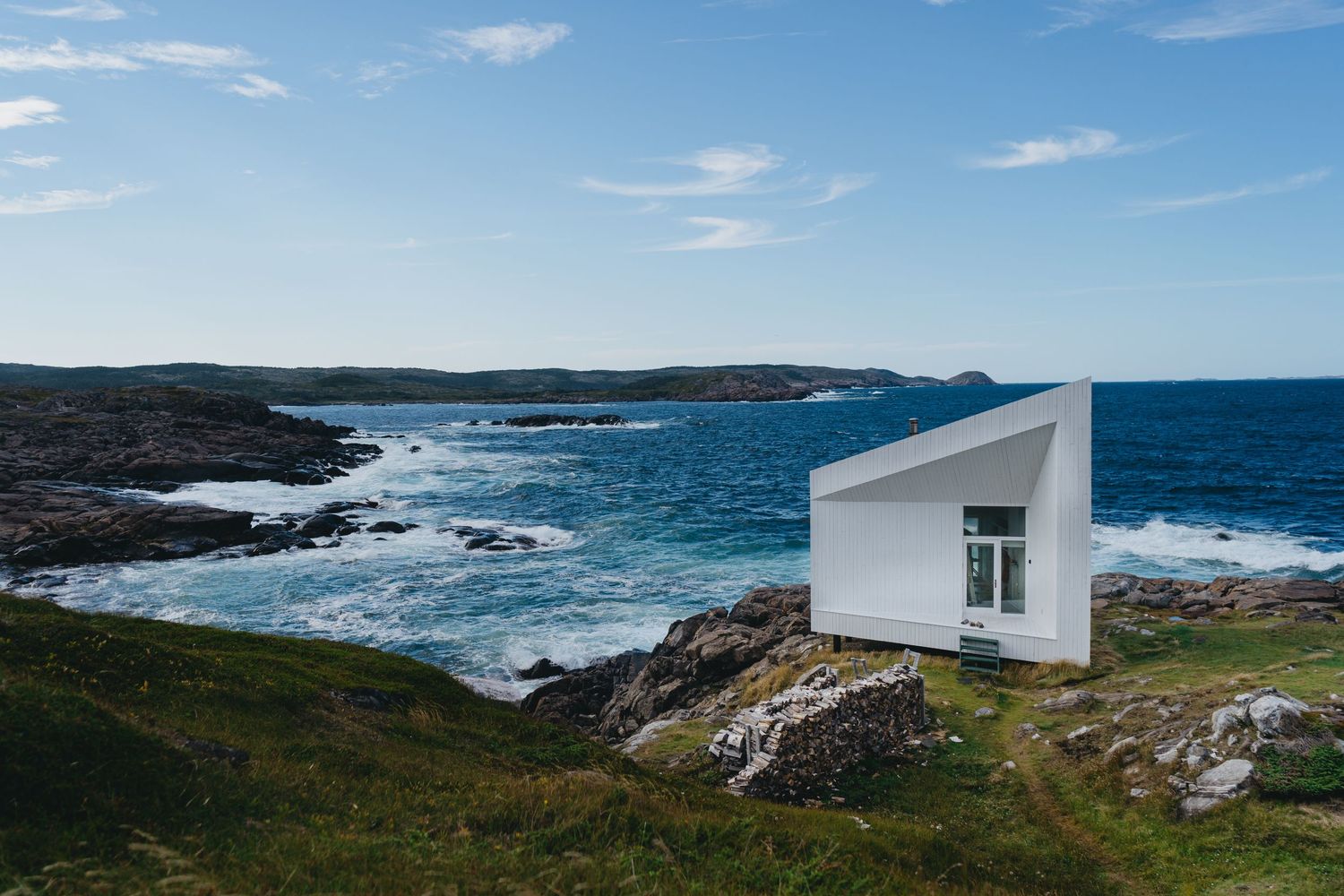 A modern white geometric building sits on rocky cliffs overlooking a dramatic ocean coastline.