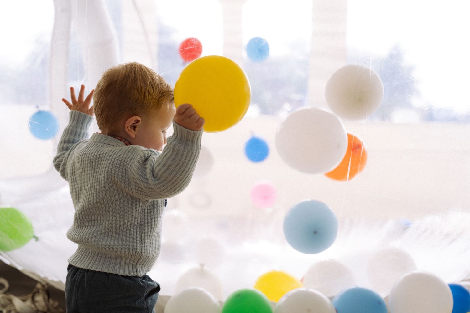 Child in gray sweater reaches up to touch colorful balloons floating on white ceiling.