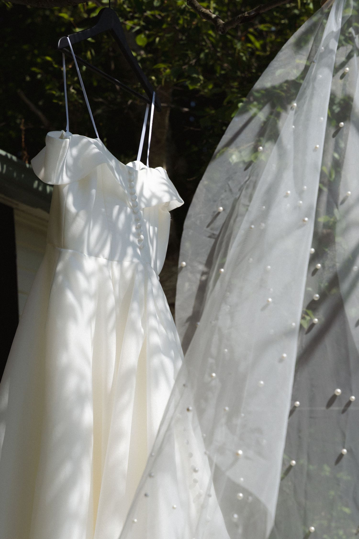 White wedding dress and veil hanging outdoors with dappled sunlight filtering through trees in the background.