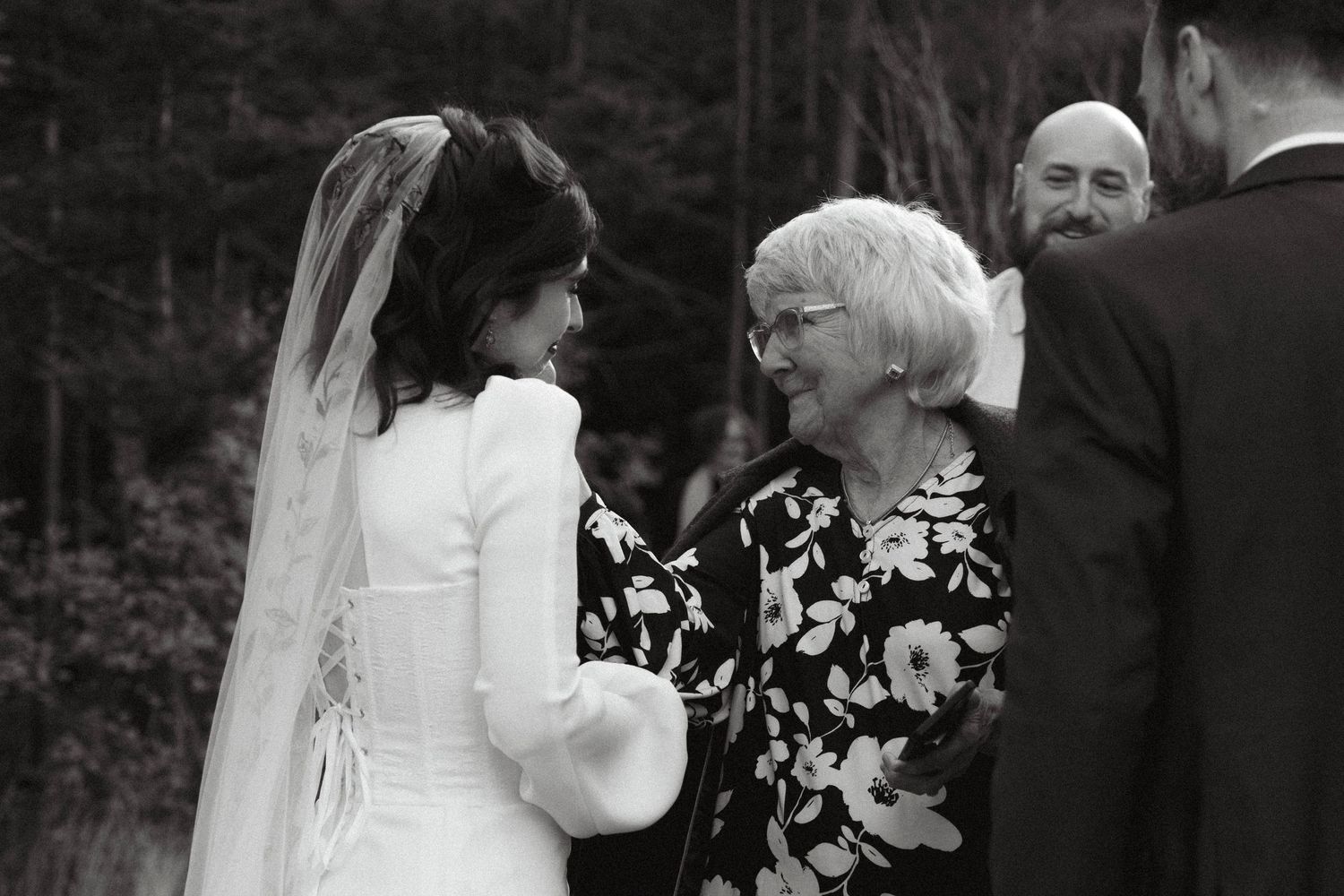 A touching moment between a bride in white and an older woman in a floral dress at an outdoor wedding ceremony.