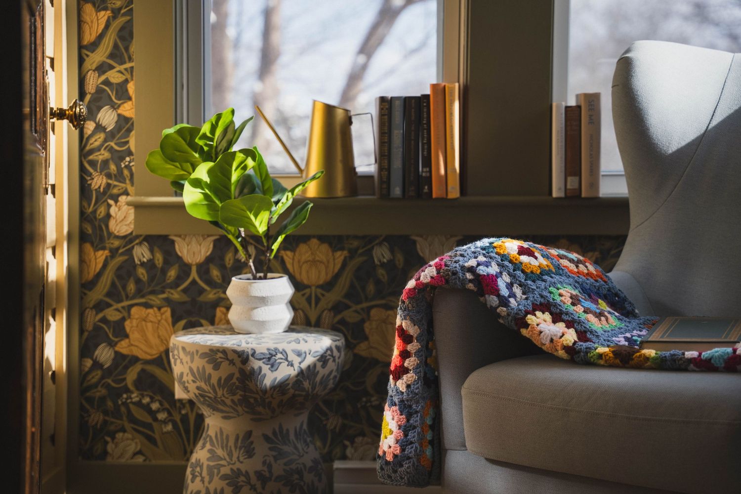 Sunlit window nook with patterned wallpaper, houseplants and reading chair.