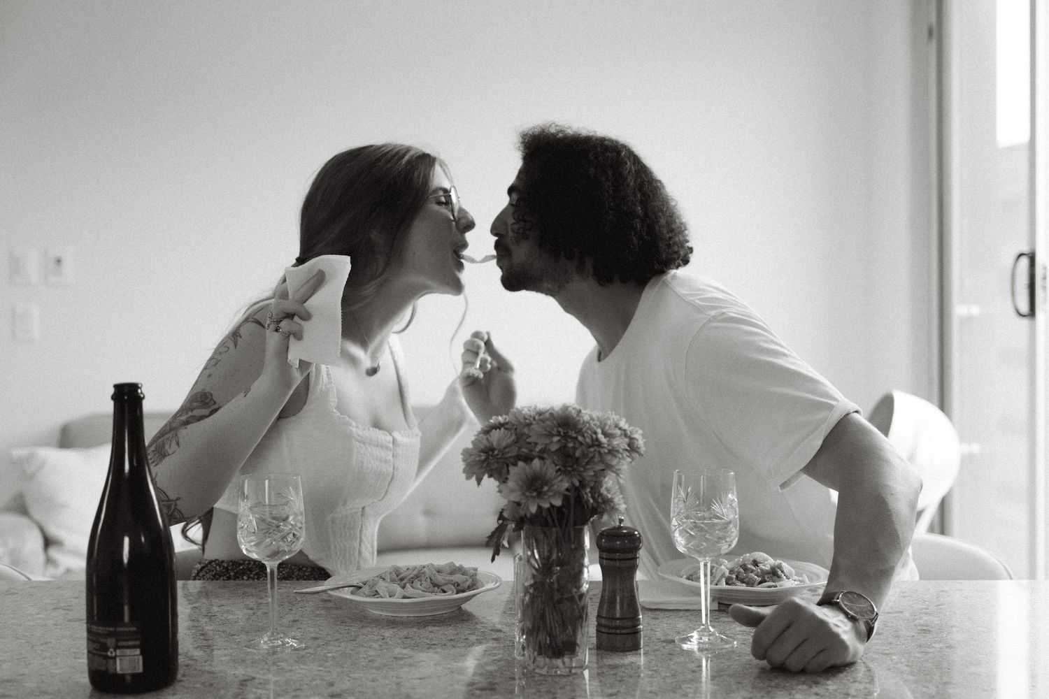 A romantic black and white scene at a dinner table with wine and a small dog between two people sharing a moment.