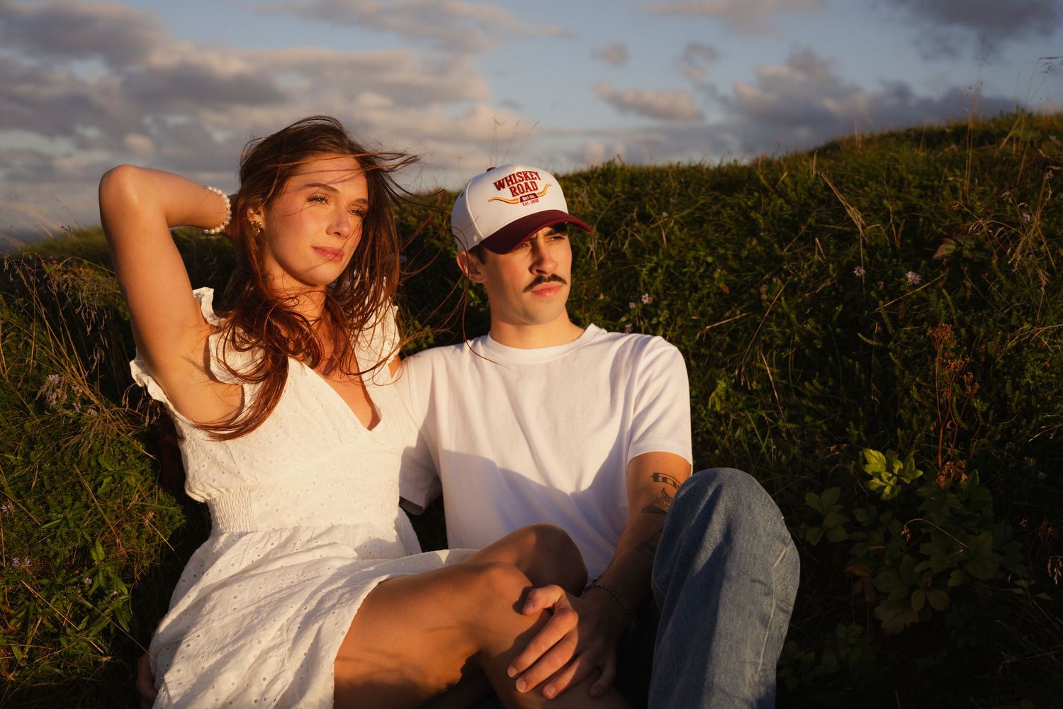 Two people relaxing in a grassy field at sunset wearing white casual clothing.
