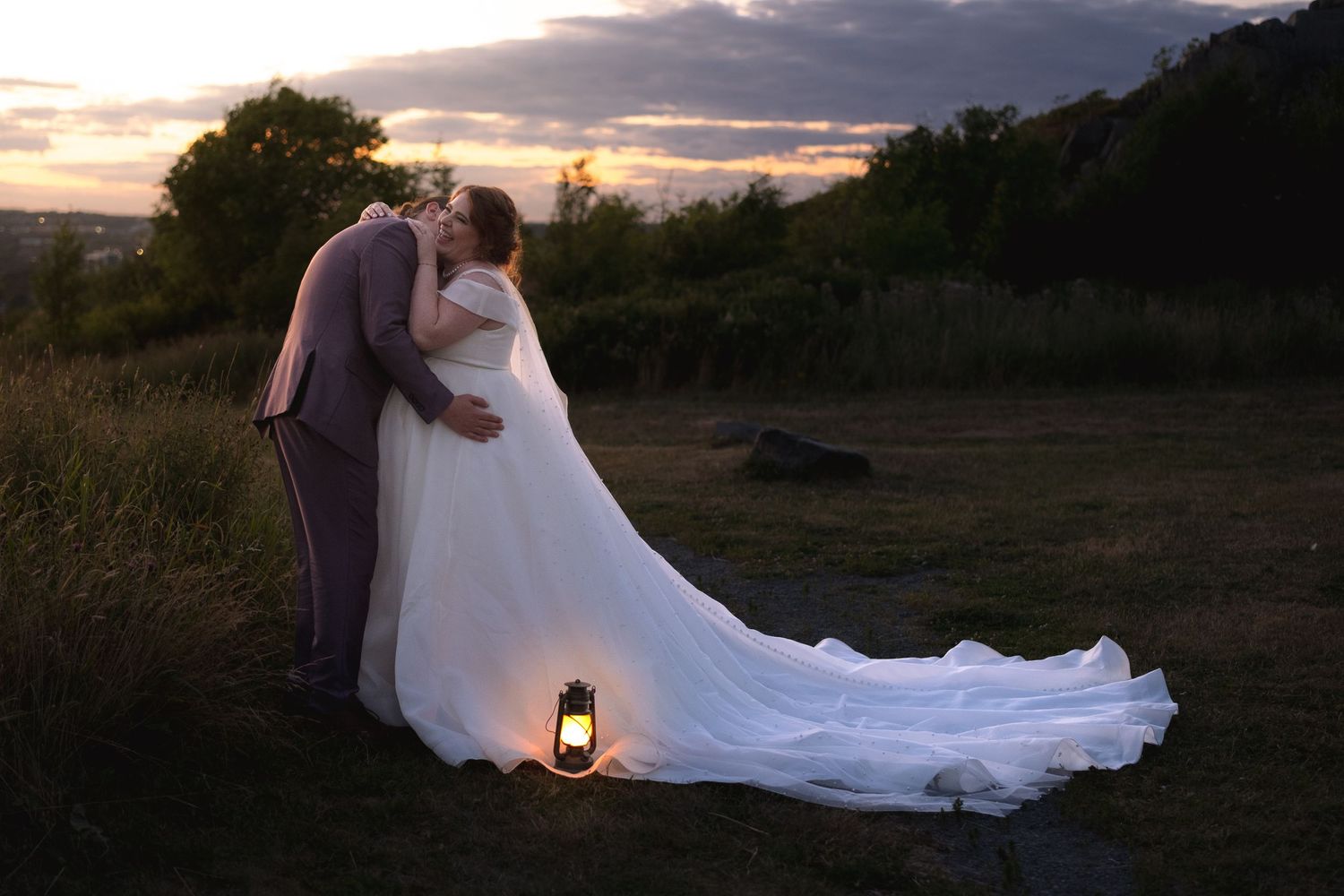 A couple in wedding attire embraces during sunset in a scenic outdoor setting as their dress flows in the breeze.