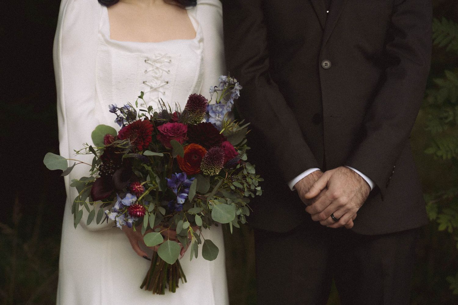Wedding couple stands together outdoors with bride holding a deep red and purple floral bouquet.