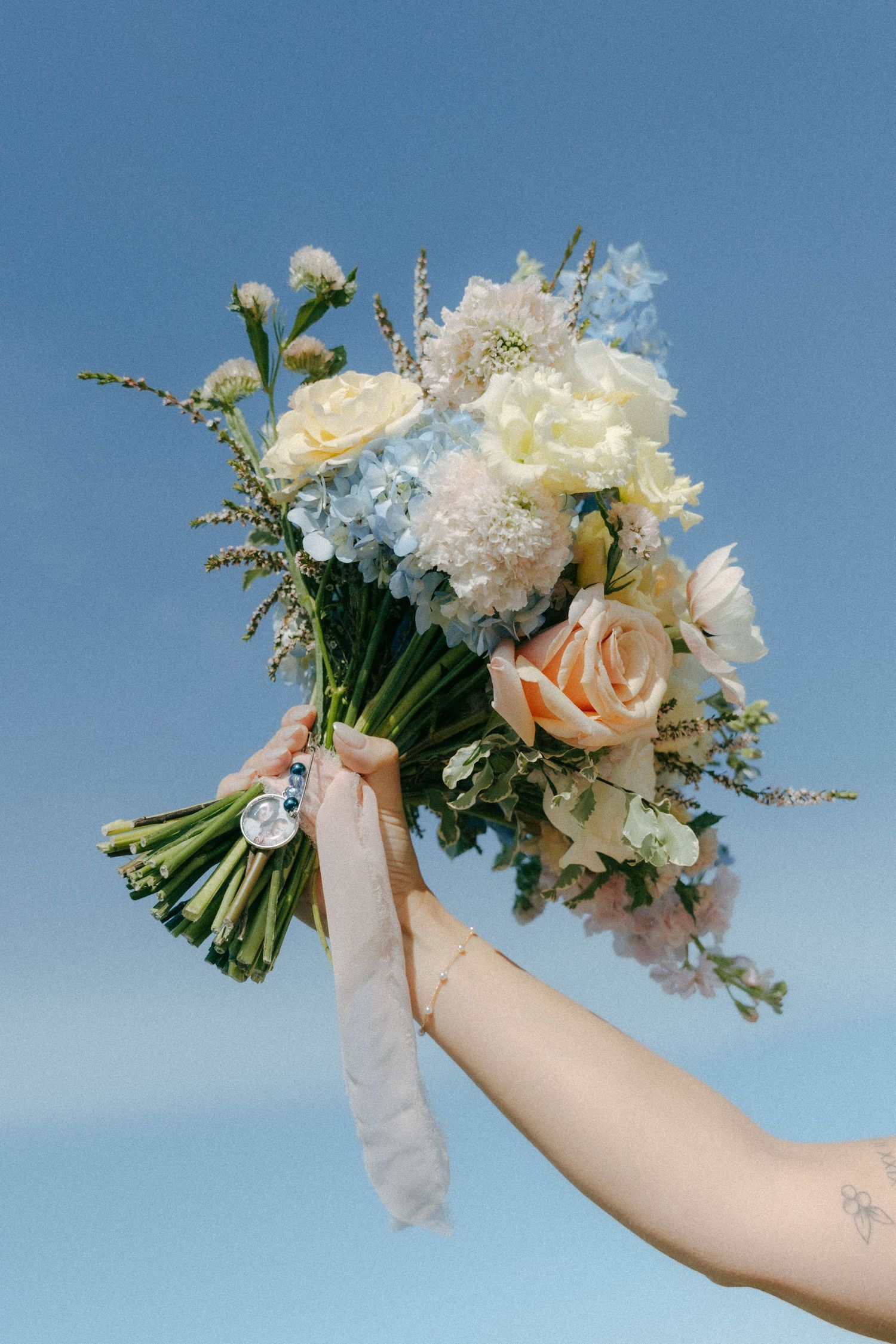 A hand holds up a romantic bouquet of white and peach roses with delicate ribbons against a bright blue sky background.