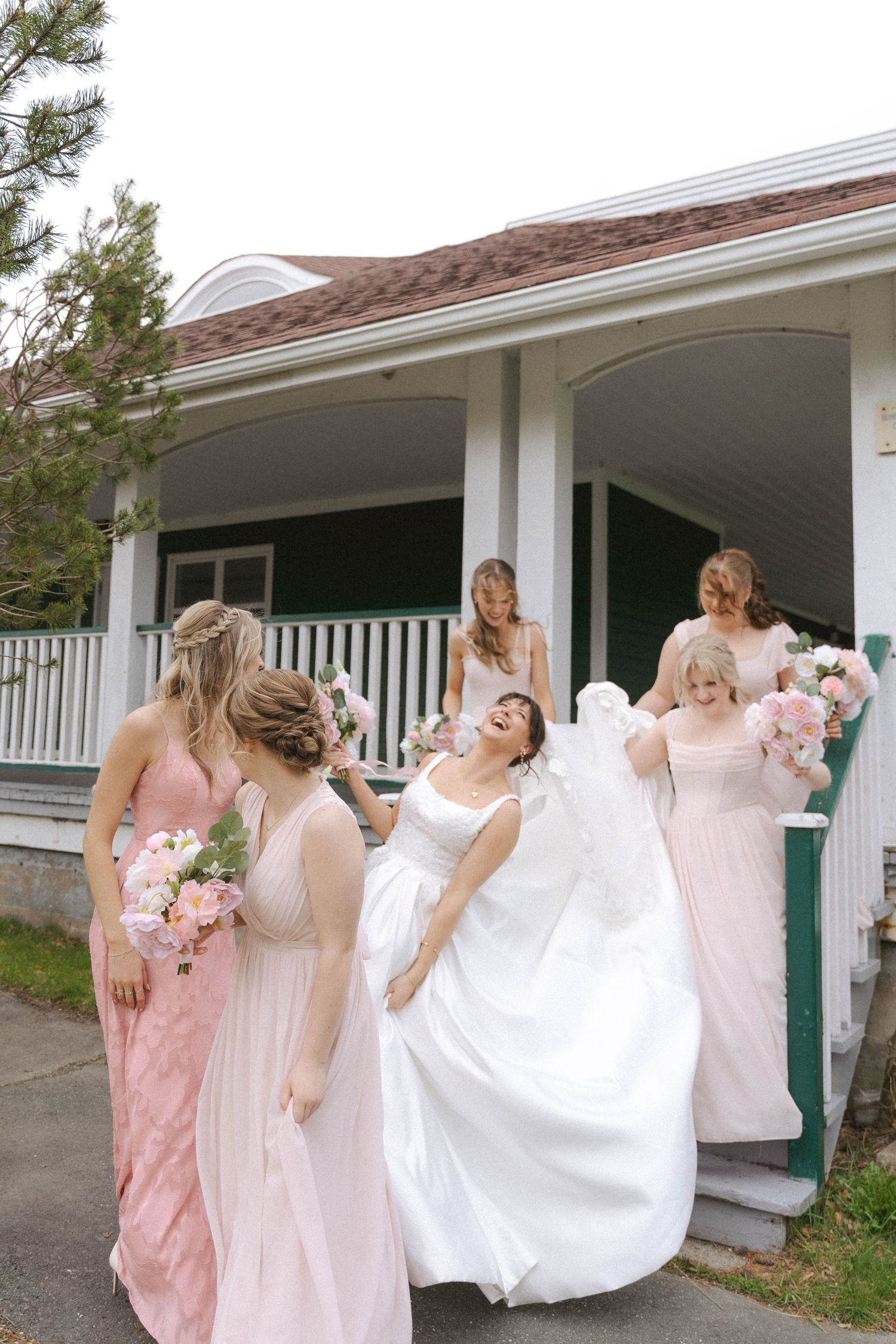 Bride in white gown laughs with bridesmaids in blush pink dresses outside a white house with a porch.