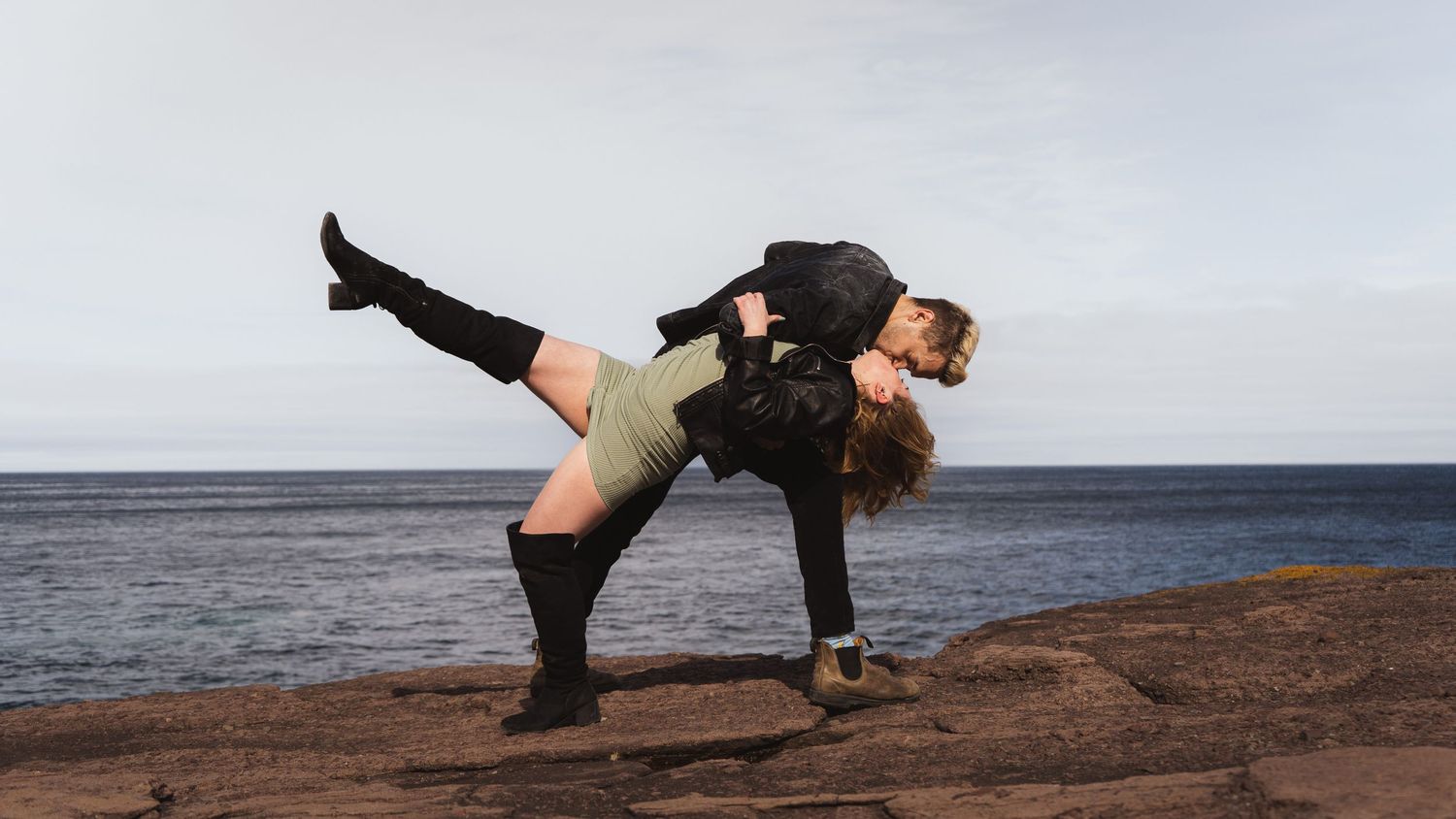 A romantic couple embraces playfully against a scenic coastal backdrop during a photoshoot.