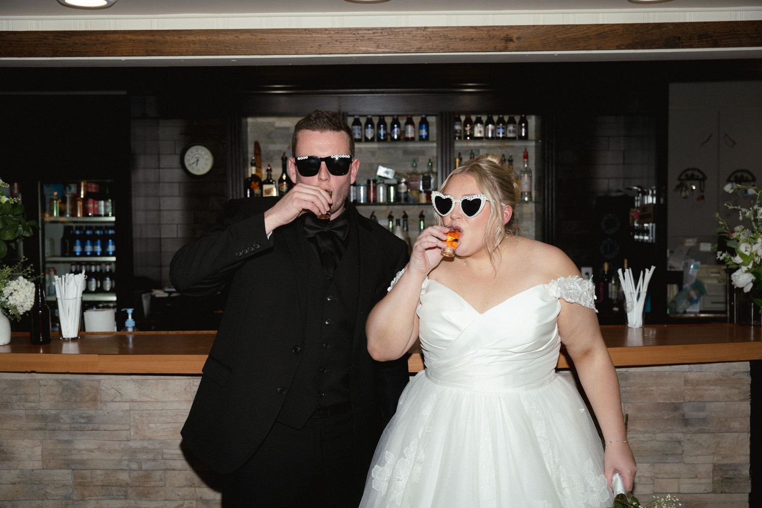 A bride in a white wedding dress holds a drink while standing next to someone in dark attire at an indoor venue.