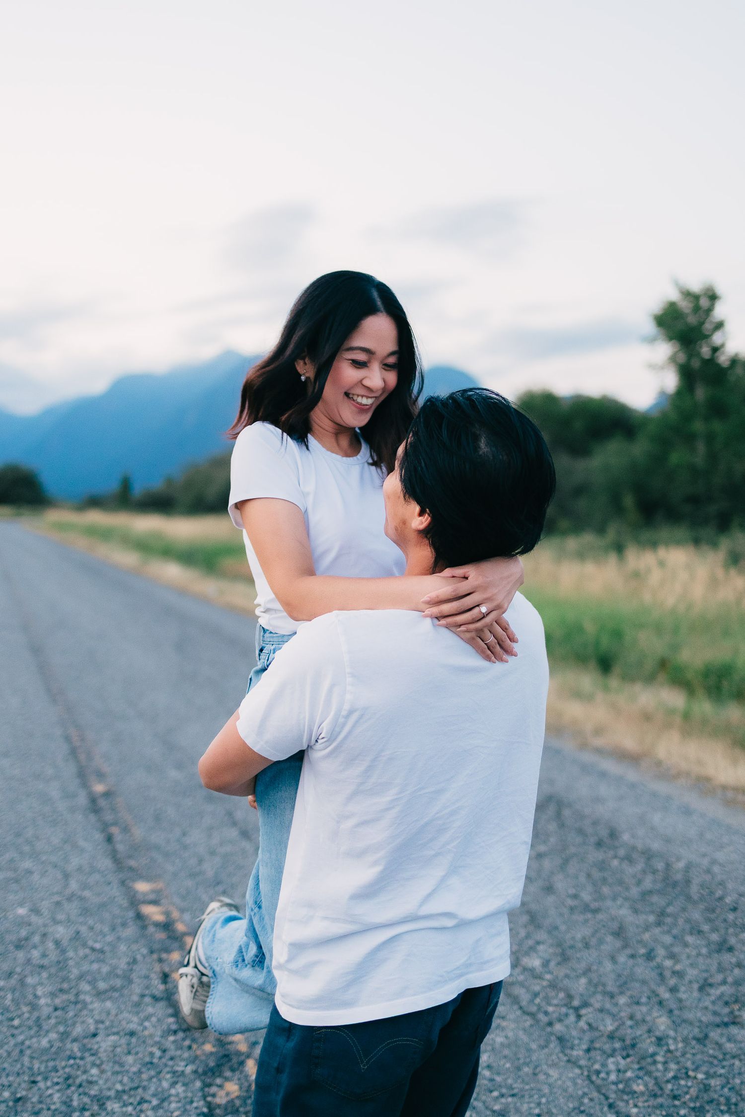 Two people in white shirts embrace on a rural road with mountains in the background.