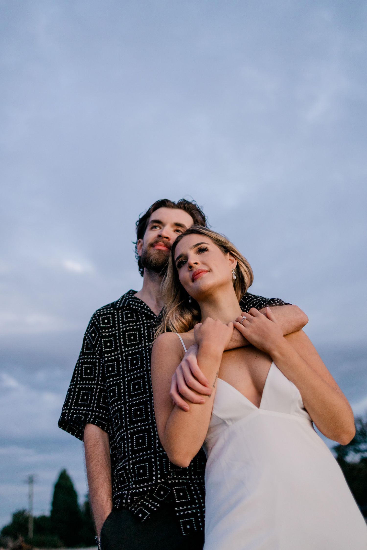 A couple in wedding attire poses dramatically against a cloudy sky during a sunset photo session.