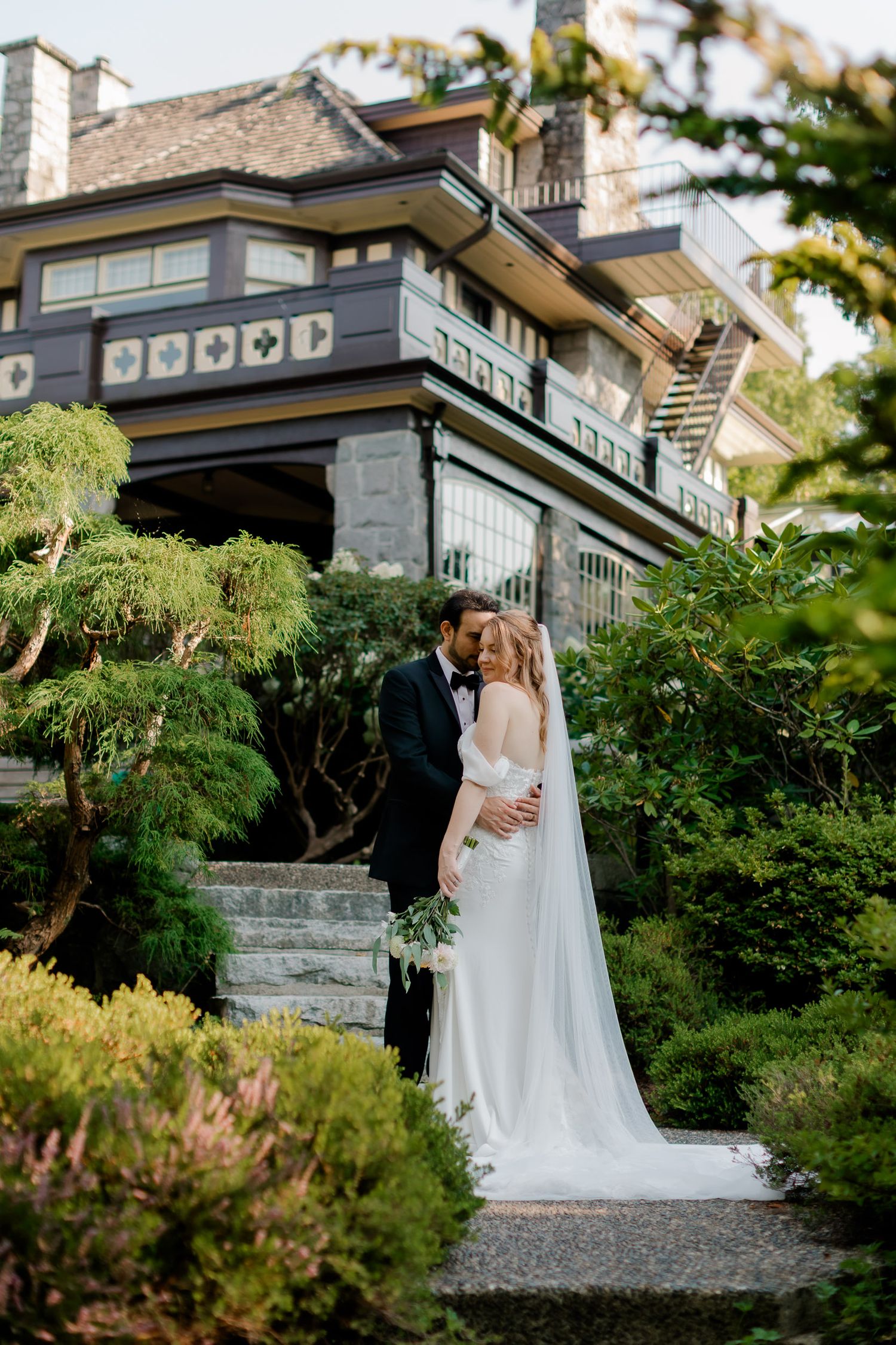 A romantic wedding couple embraces on stone steps surrounded by lush greenery at an elegant venue at sunset.