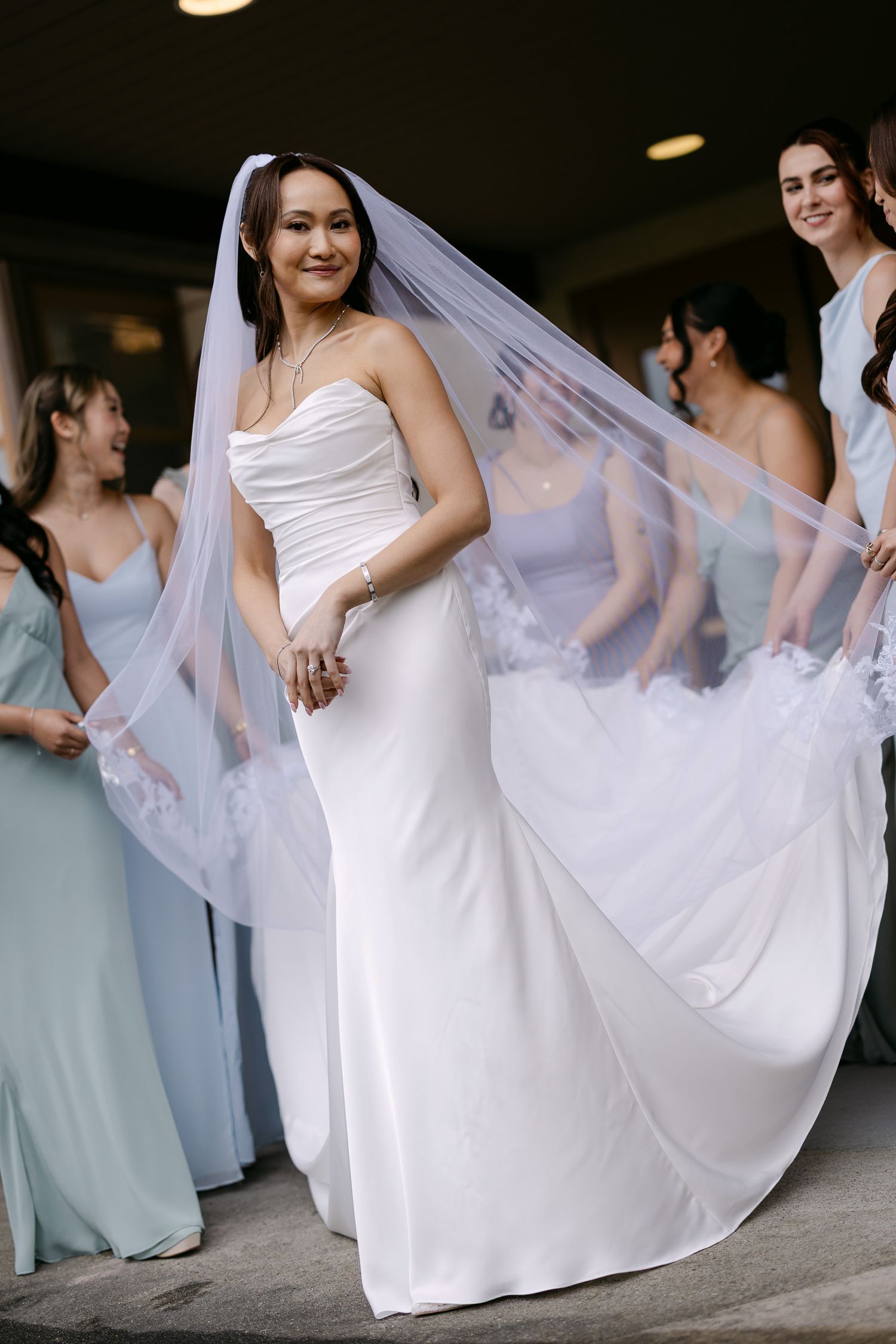Bride in white strapless gown and long veil held by bridesmaids in light blue dresses outdoors.