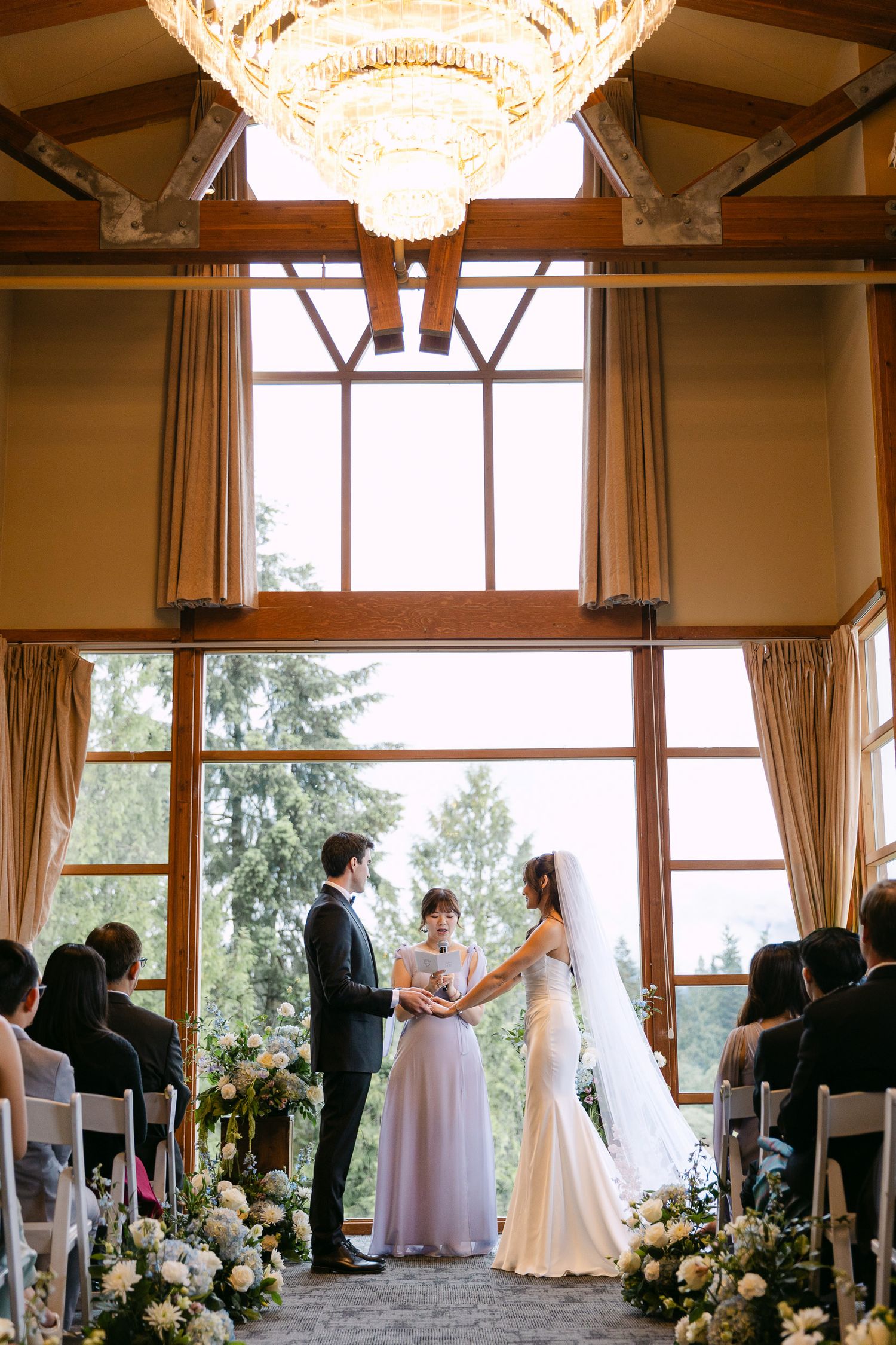 Elegant indoor wedding ceremony with couple exchanging vows before large windows, floral arrangements, and a grand chandelier overhead.