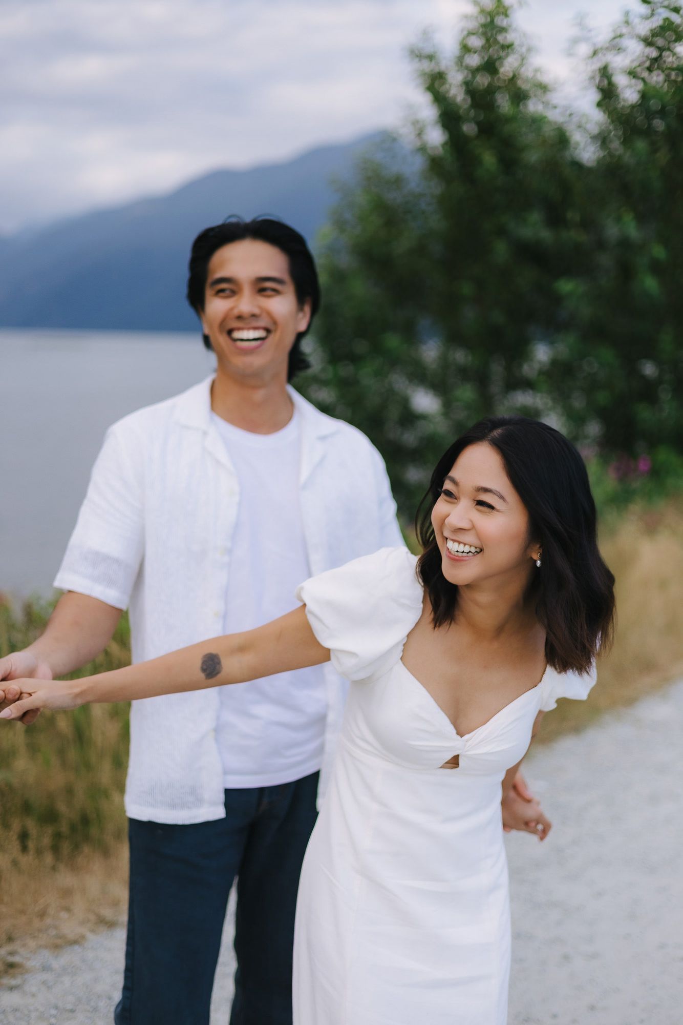 Laughing couple in white outfits play by a mountain lake during a sunset engagement photo shoot.