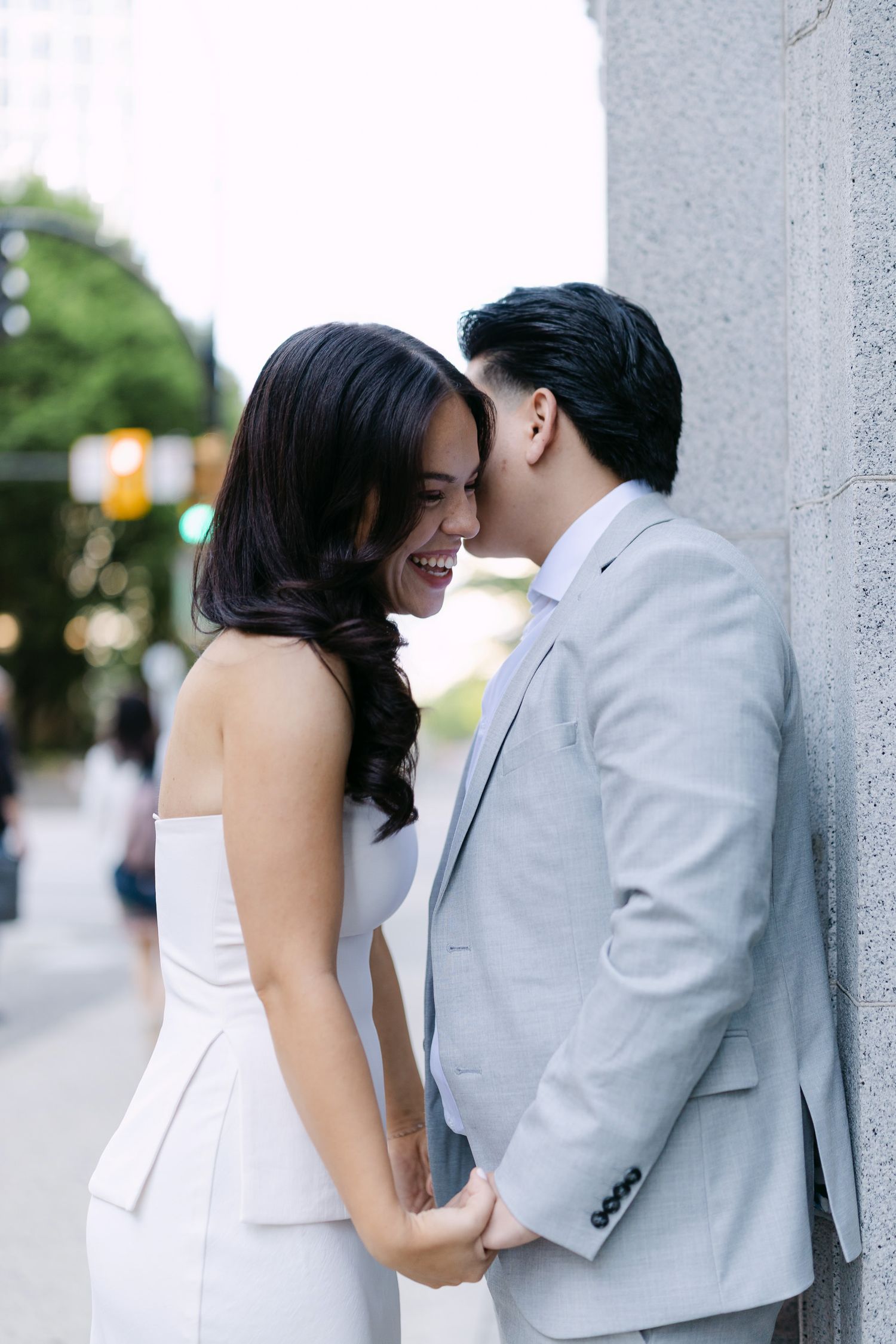 Close-up engagement photo of a couple holding hands as a man in a gray suit kisses the cheek of a laughing woman in white.