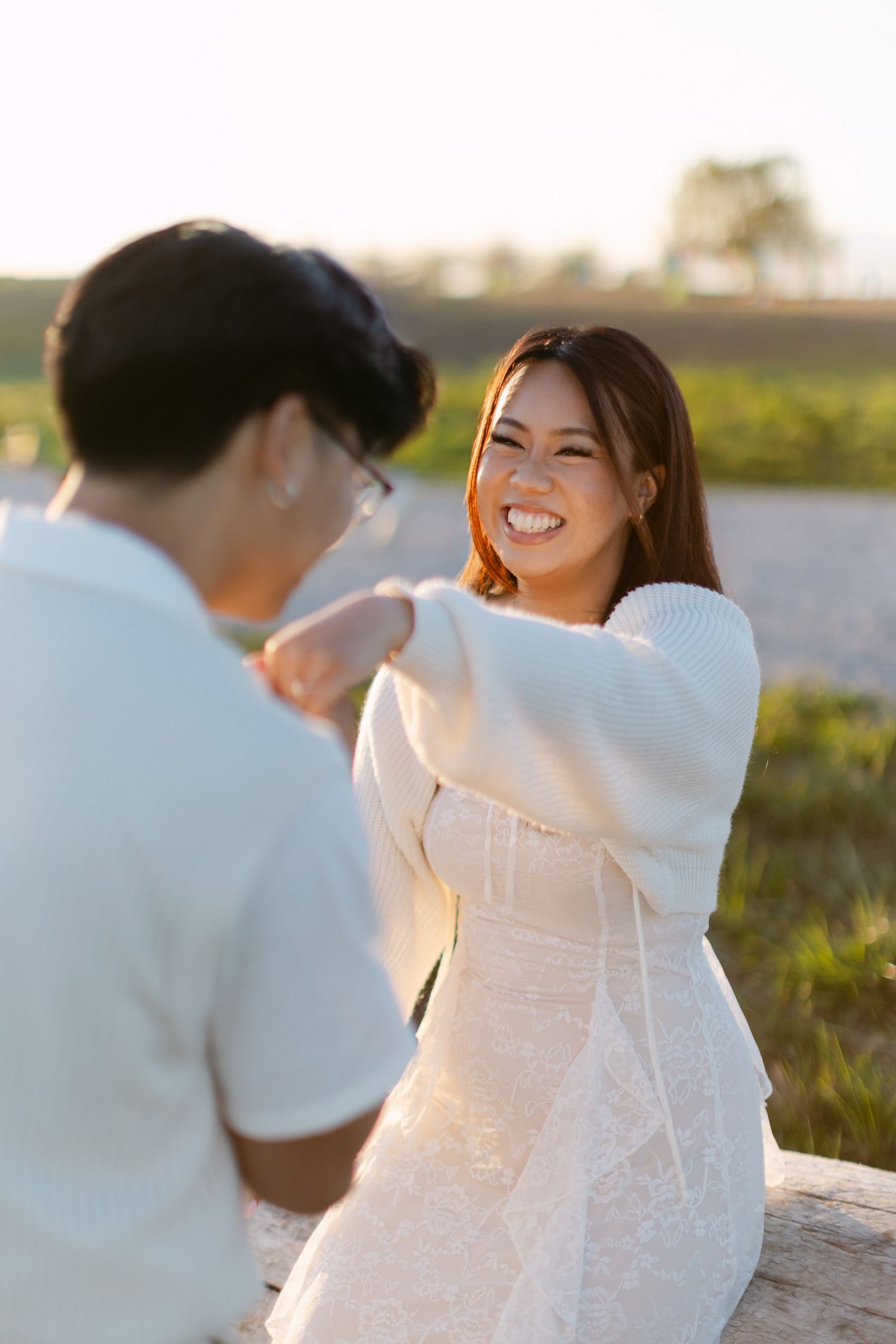 Joyful woman in a white lace dress laughing while playfully reaching toward her fiancé on a log at sunset.