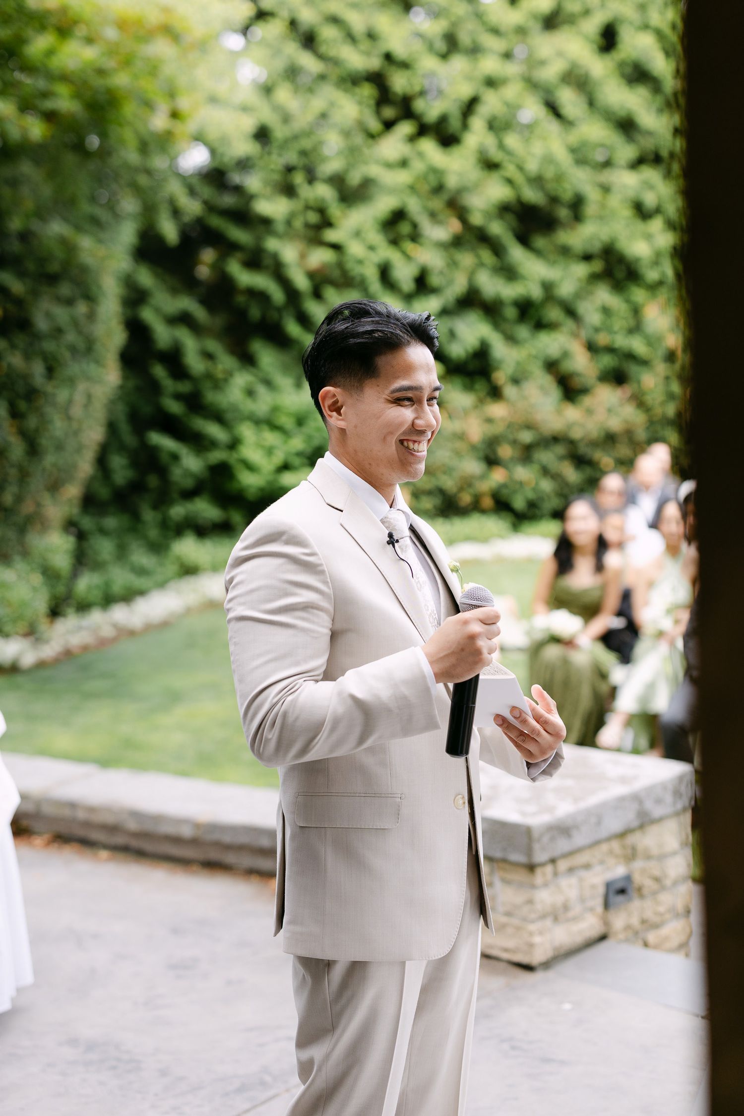Smiling groom in beige suit holding microphone at outdoor wedding ceremony with lush green hedges.