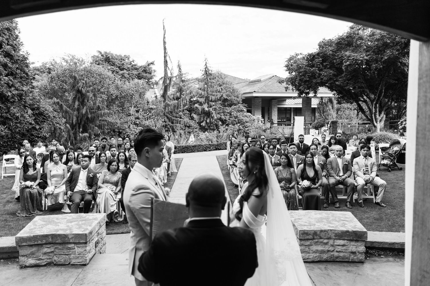 Black and white outdoor wedding ceremony with couple at altar and guests seated in garden.