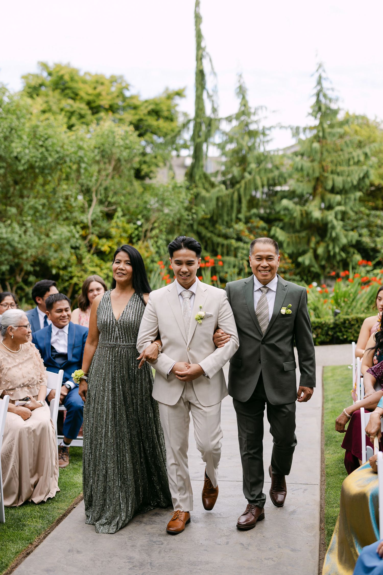Groom in ivory suit walks down outdoor aisle escorted by parents at a garden wedding ceremony.
