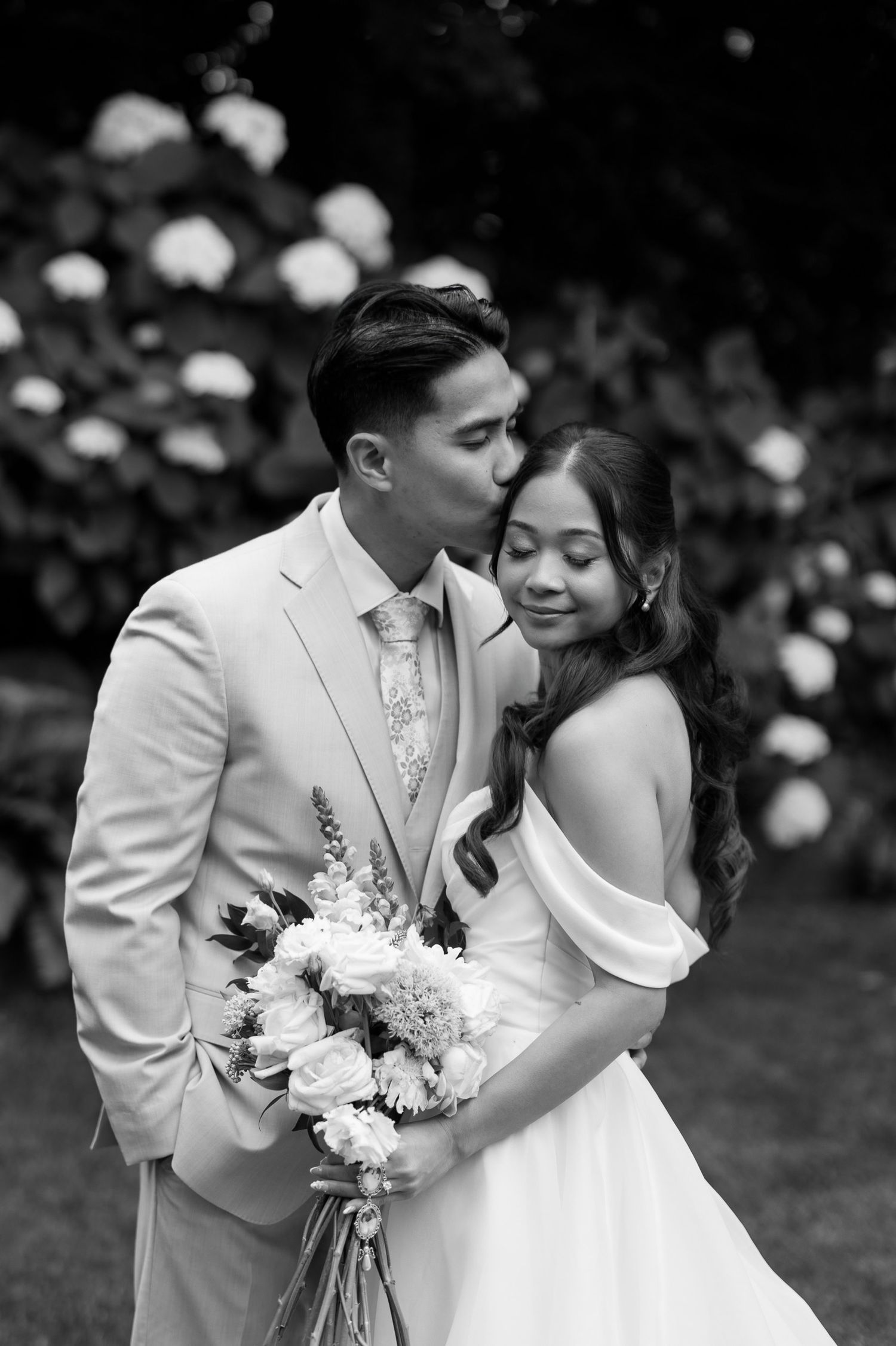 Black and white wedding photo of groom kissing bride on cheek while she smiles holding a bouquet outdoors.